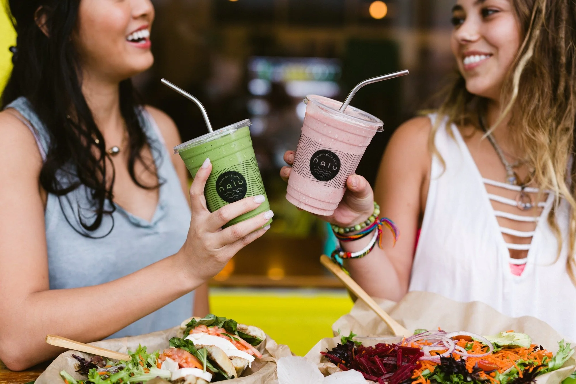 Two women smiling and enjoying drinks at a restaurant, holding colorful smoothies; one green and one pink, with food like salads and sandwiches on the table.