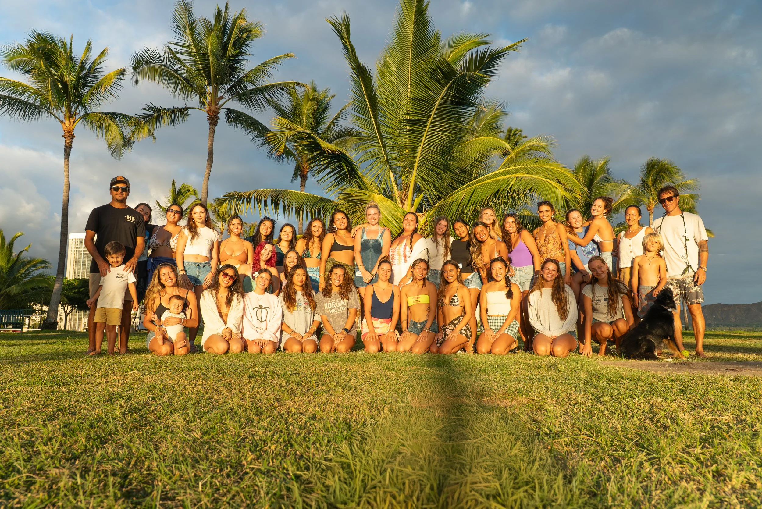 Group of people, including children and adults, posing outdoors on a grassy area with palm trees and a cloudy sky in the background during sunset.