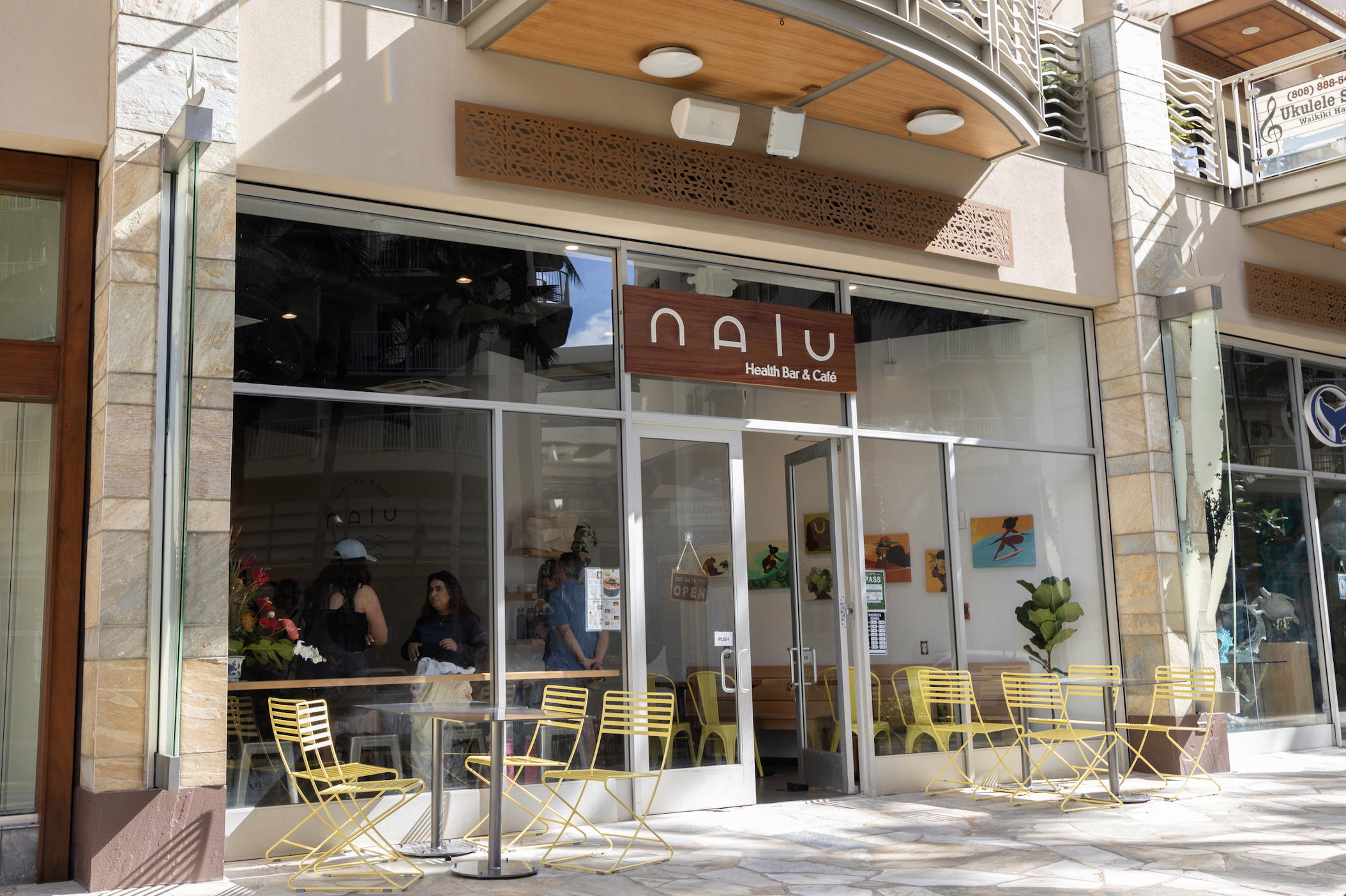 storefront of NAU Health Bar & Cafe with yellow outdoor chairs and large glass windows, inside three women are talking, decor includes paintings and potted plants, sunny day.