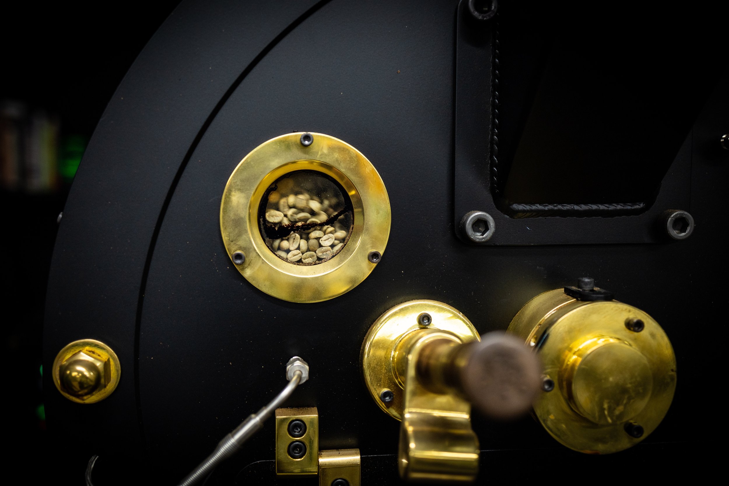 Close-up of a black and gold coffee roasting machine with green coffee beans inside a viewing window.