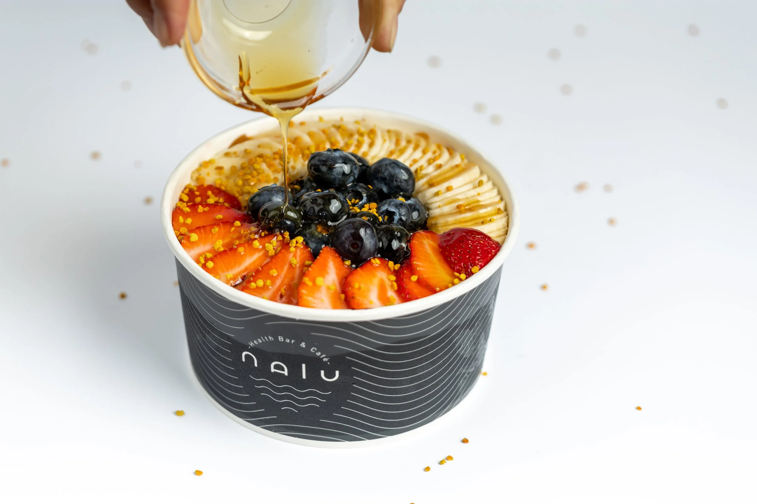 A cup of yogurt topped with strawberries, blueberries, and chopped nuts, with honey being drizzled on top.