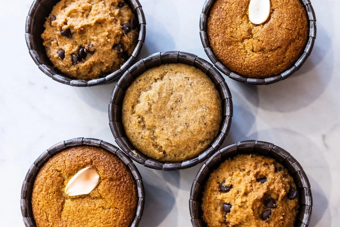 Five muffins in dark woven baskets on a light surface, some topped with almonds and some with chocolate chips.