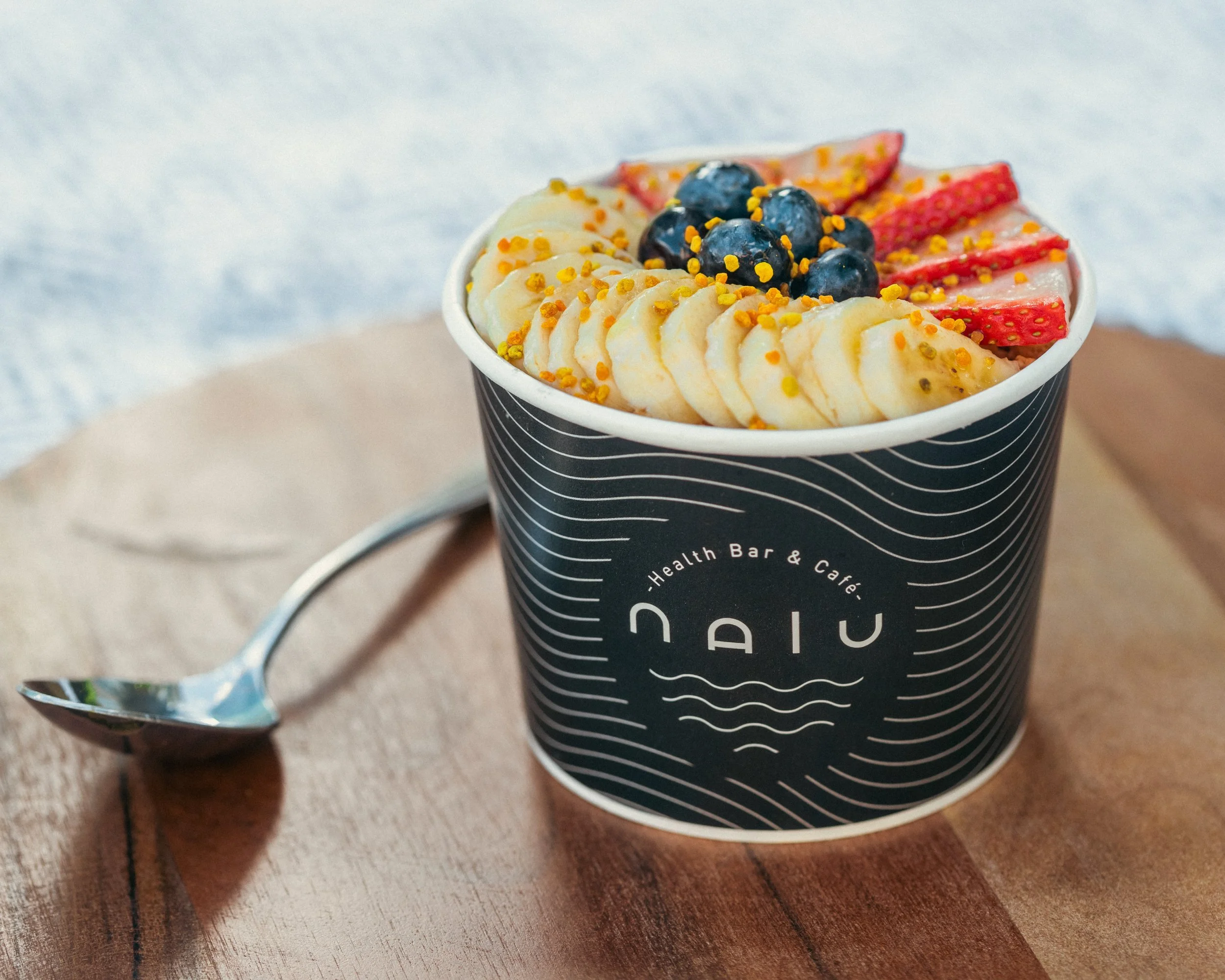 A cup of smoothie topped with banana slices, blueberries, strawberry slices, and sprinkled with yellow topping, placed on a wooden surface with a spoon beside it.