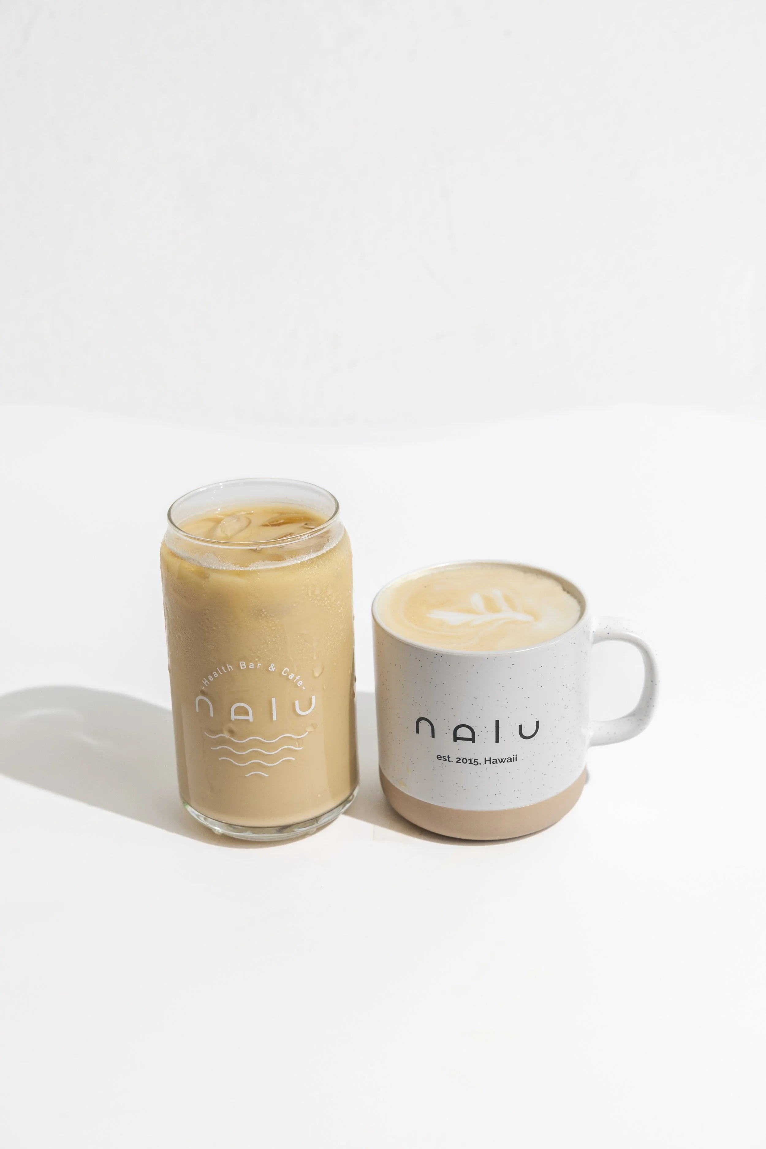 A person holding a white coffee mug with black text that reads 'NADIU' and 'Health Bar & Cafe' in front of a yellow background, with a latte inside showing latte art on top.