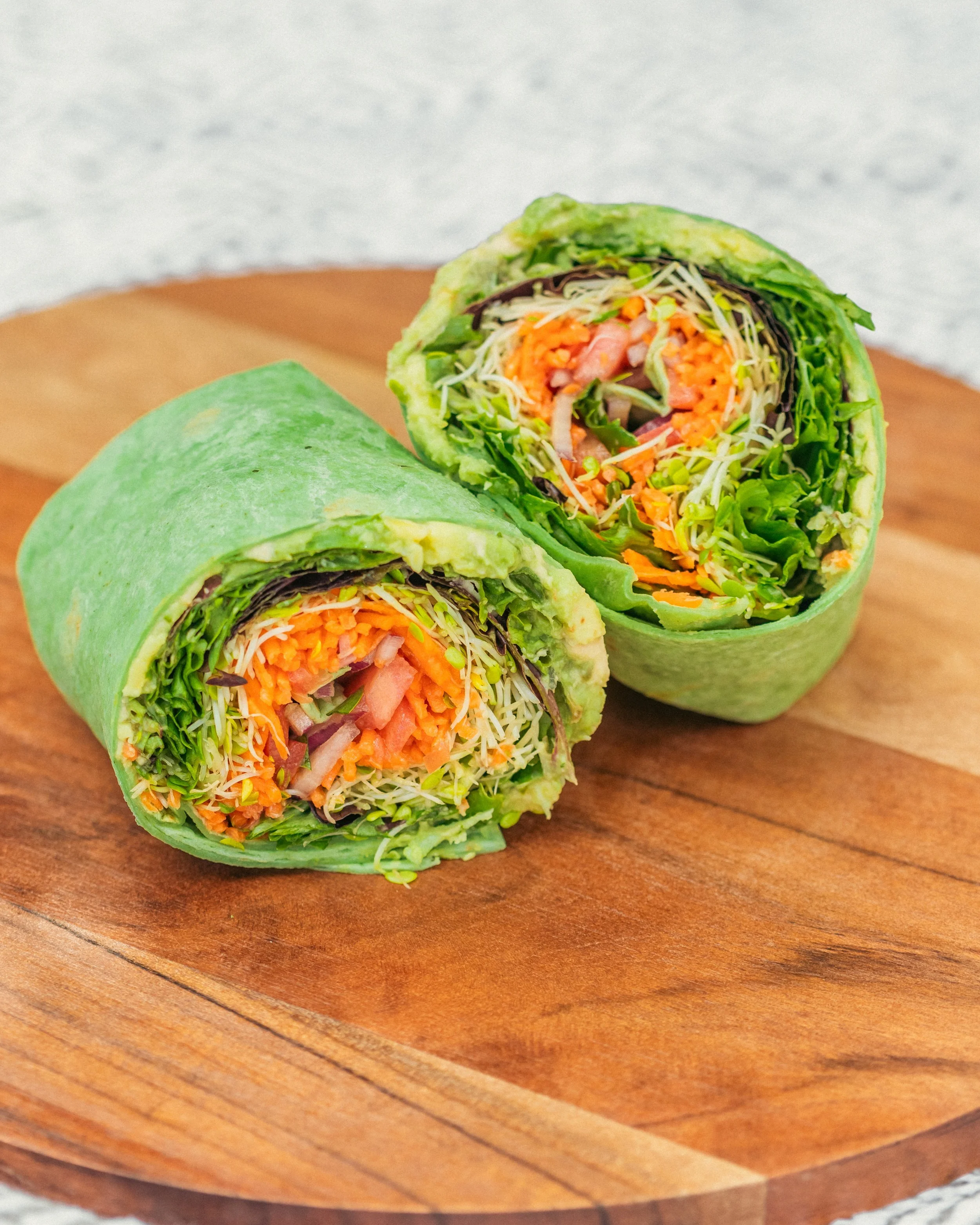 A close-up of two veggie wraps cut in half, showing lettuce, shredded carrots, sprouts, and other vegetables, on a wooden plate.