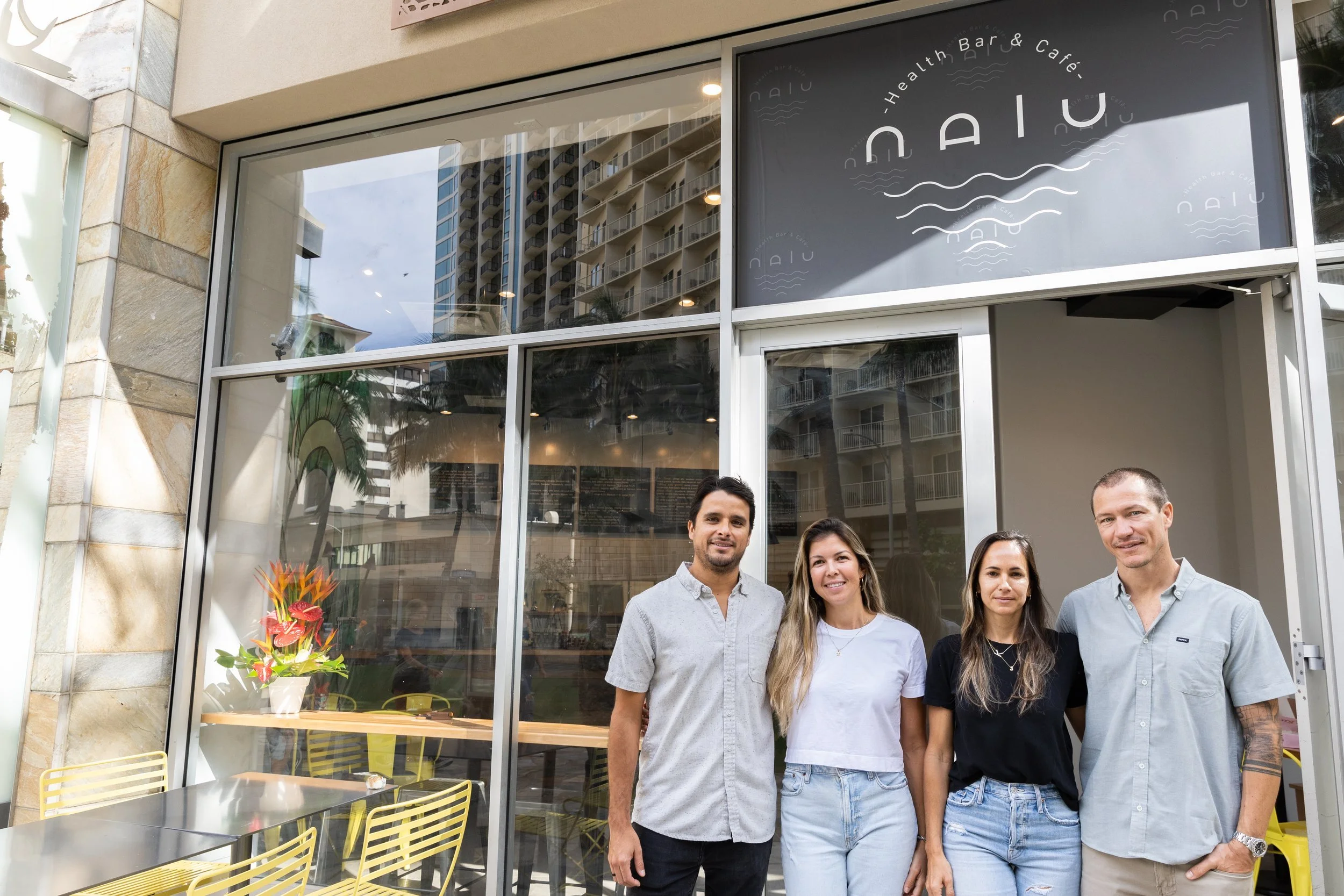 Four people standing outside a restaurant called NAIU, with a glass storefront, a potted plant with flowers on a table inside, and a cityscape of tall buildings reflected in the windows.