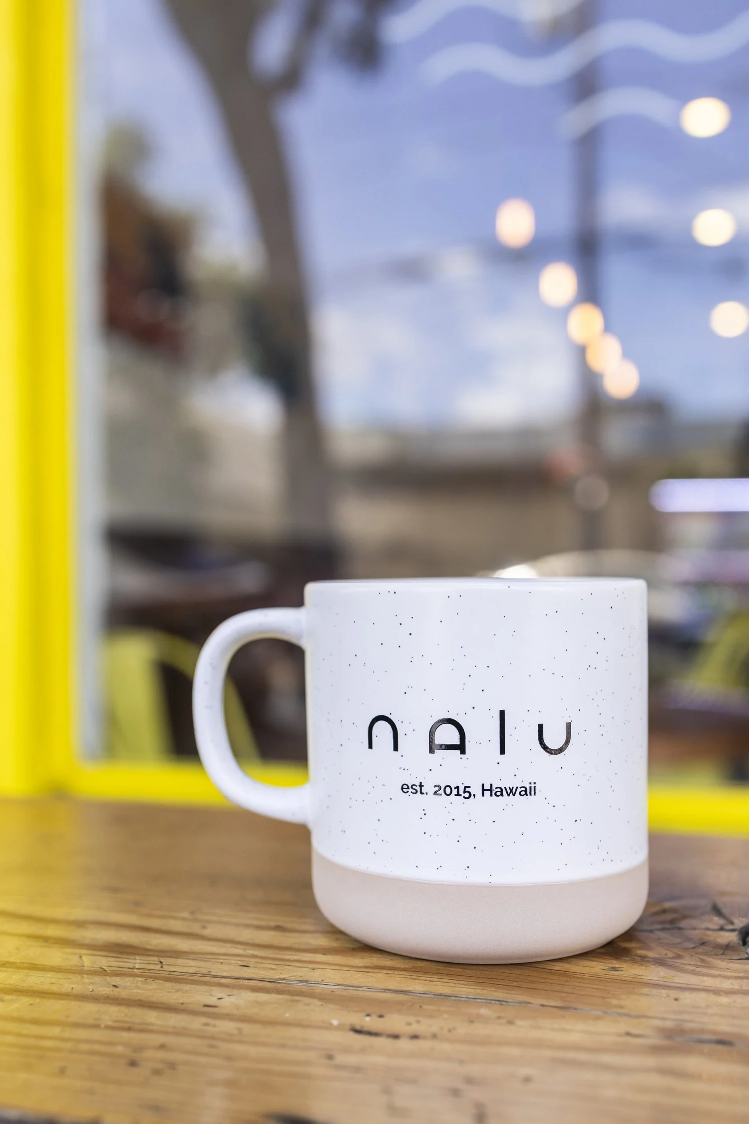 A white speckled mug with the word 'nalu' and 'est. 2015, Hawaii' printed on it, placed on a wooden table near a yellow window frame, with blurred outdoor background and string lights.