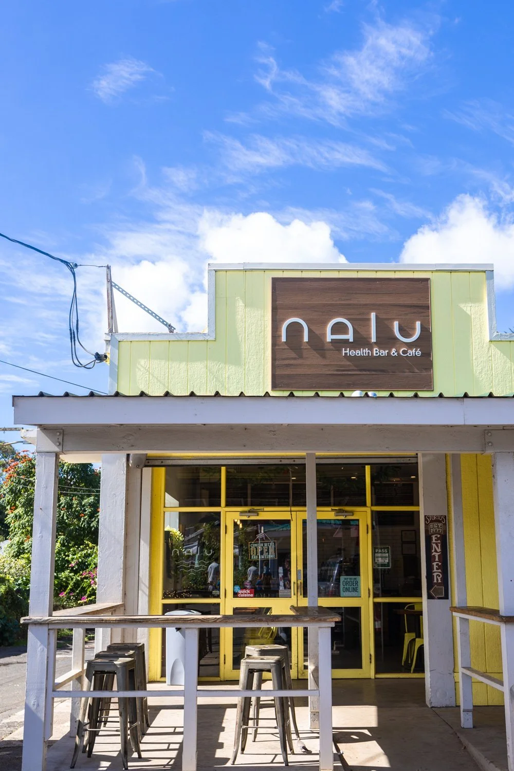 Front view of a building with yellow and white exterior, sign reads 'Nalu Health Bar & Café,' with outdoor seating and clear blue sky.