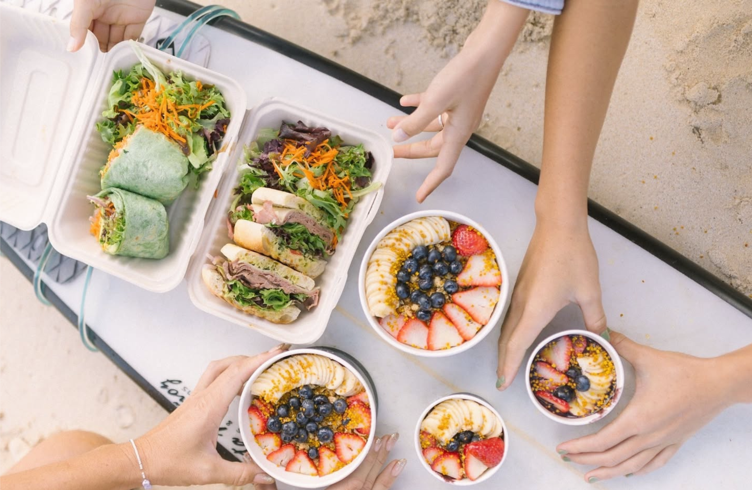 People sharing colorful fruit bowls, salads, and wraps on a picnic table at the beach.