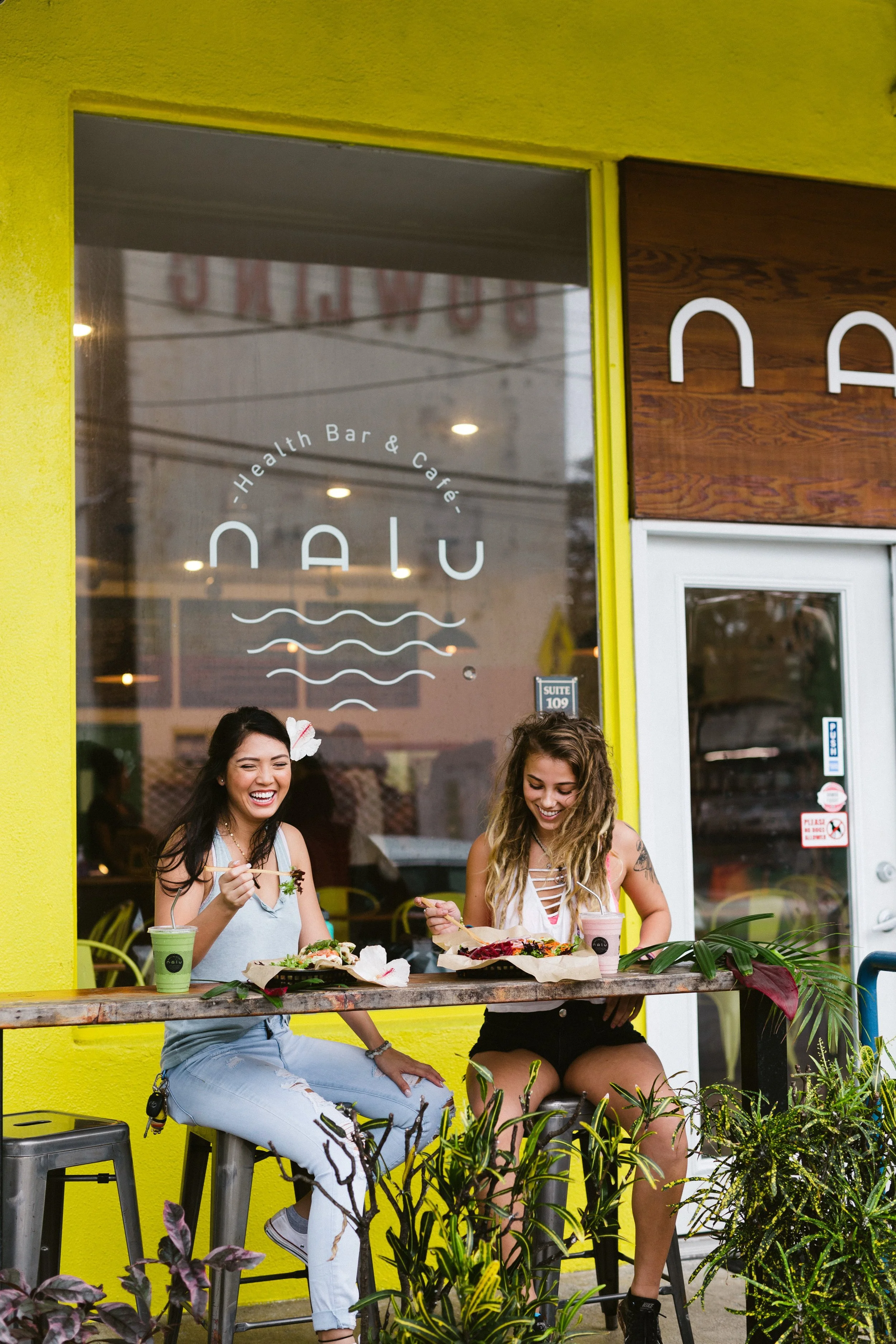 Two women sitting outside a cafe, laughing and eating with drinks. The cafe has a neon sign and a yellow exterior wall.