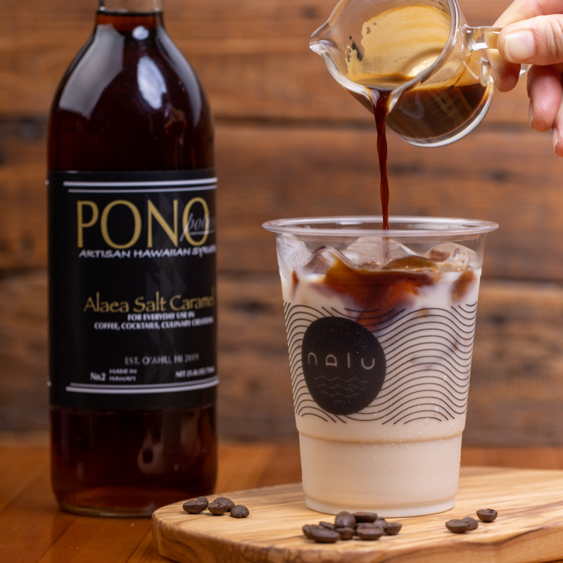 Pouring coffee syrup into a glass of iced coffee with ice cubes and milk, with a bottle of PONO Alaea Salt Caramel syrup and some coffee beans on a wooden surface.