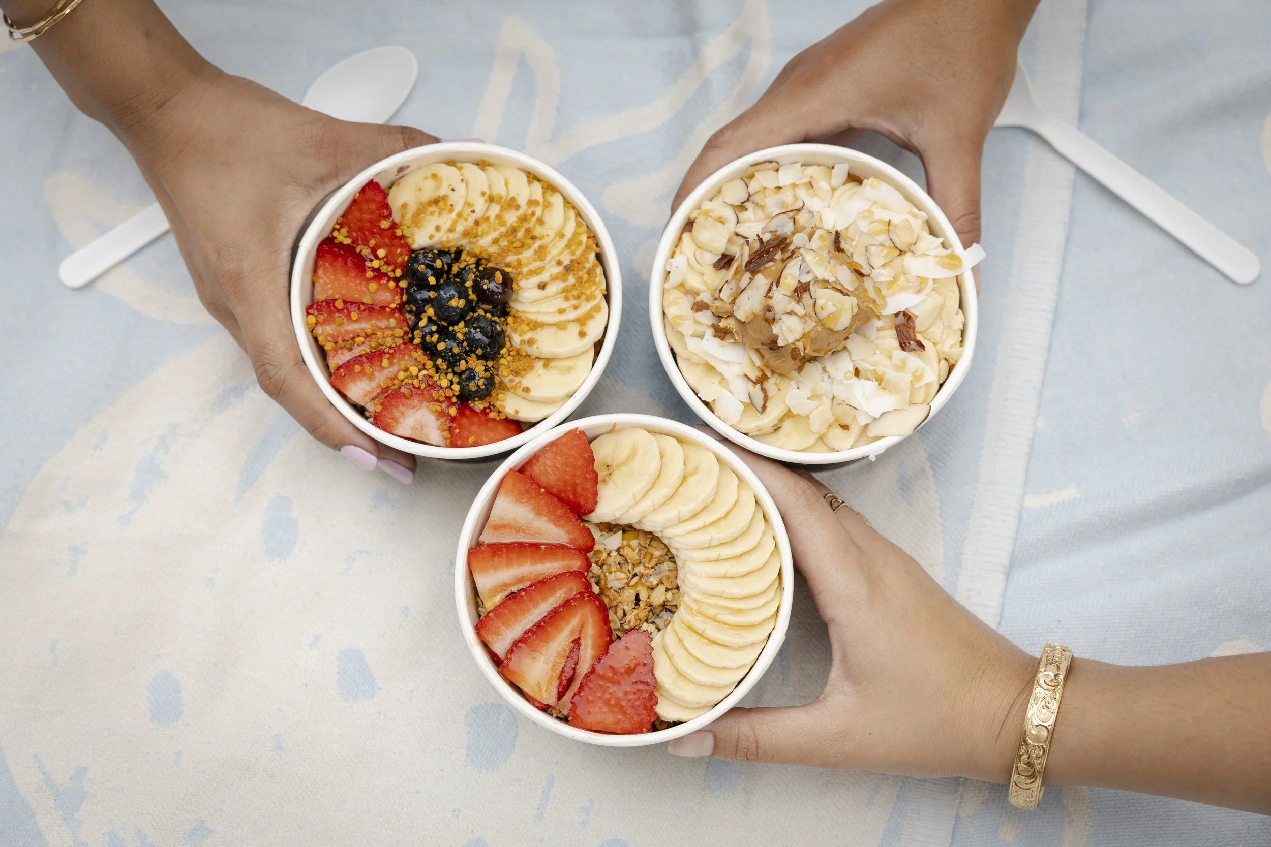 Three cups of frozen yogurt with various toppings being held by three different hands over a light-colored table.