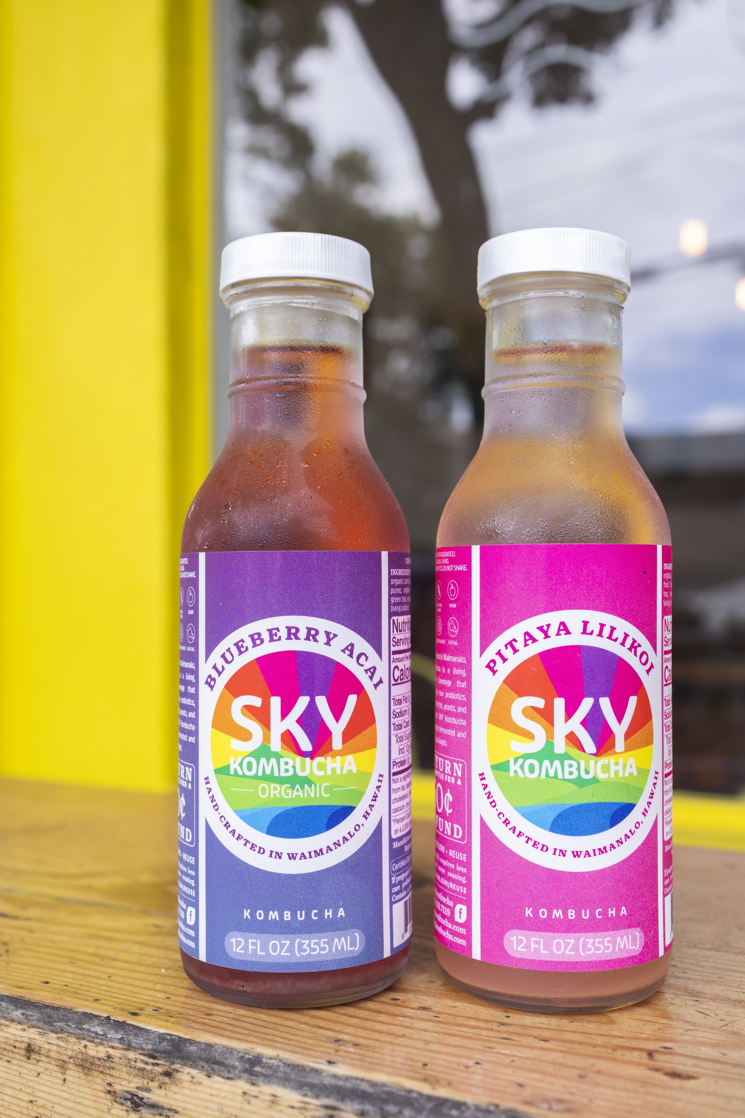 Two bottles of Sky Kombucha, one with a purple label and the other with a pink label, sitting on a wooden surface outdoors. The labels display flavors, blueberry acai and pitaya lilikon, with rainbow-colored sky and mountain graphics.