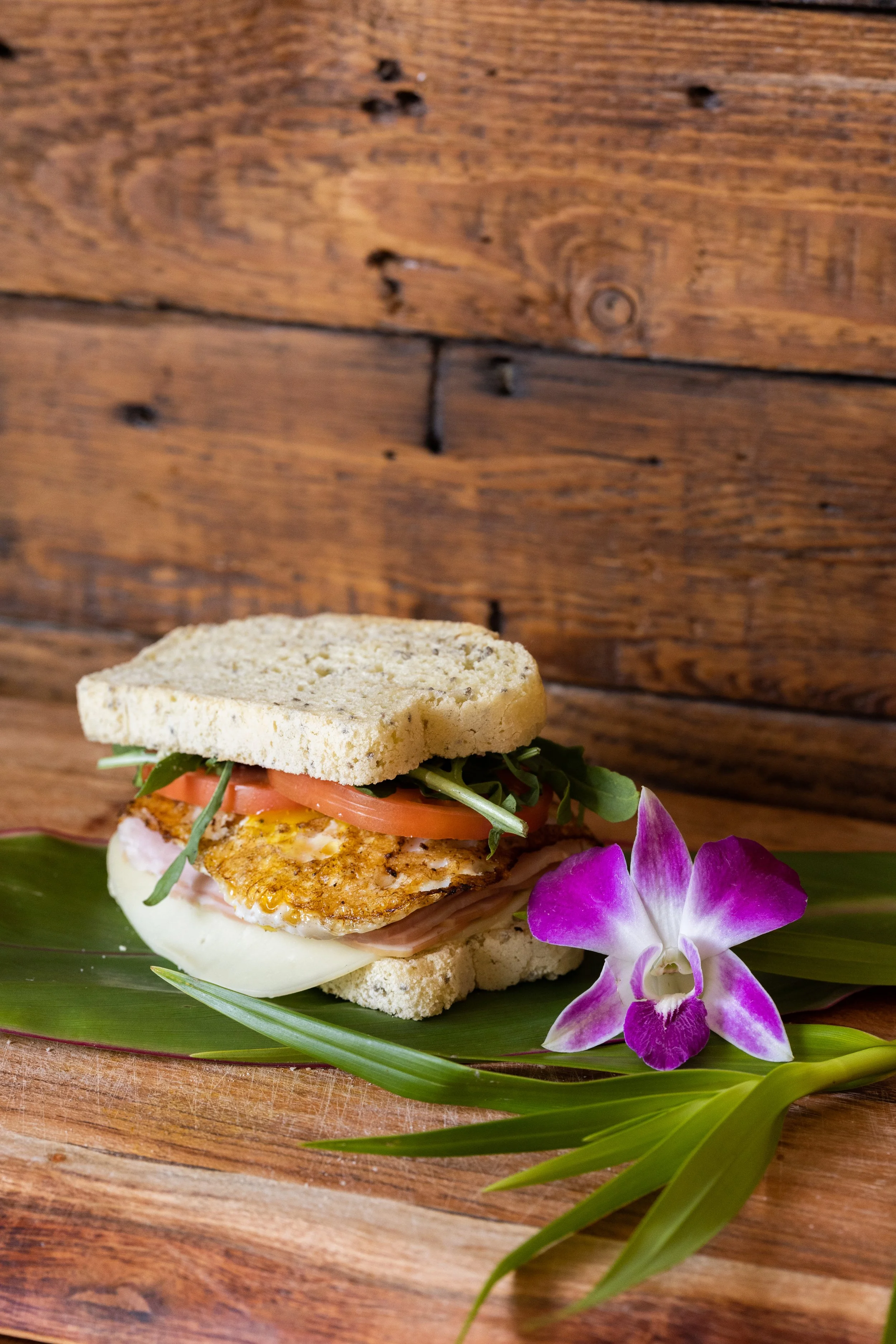 Sandwich with fried egg, tomato, greens, and turkey on white bread, garnished with pink and purple orchid and green leaves, on a green leaf placed on a wooden surface.