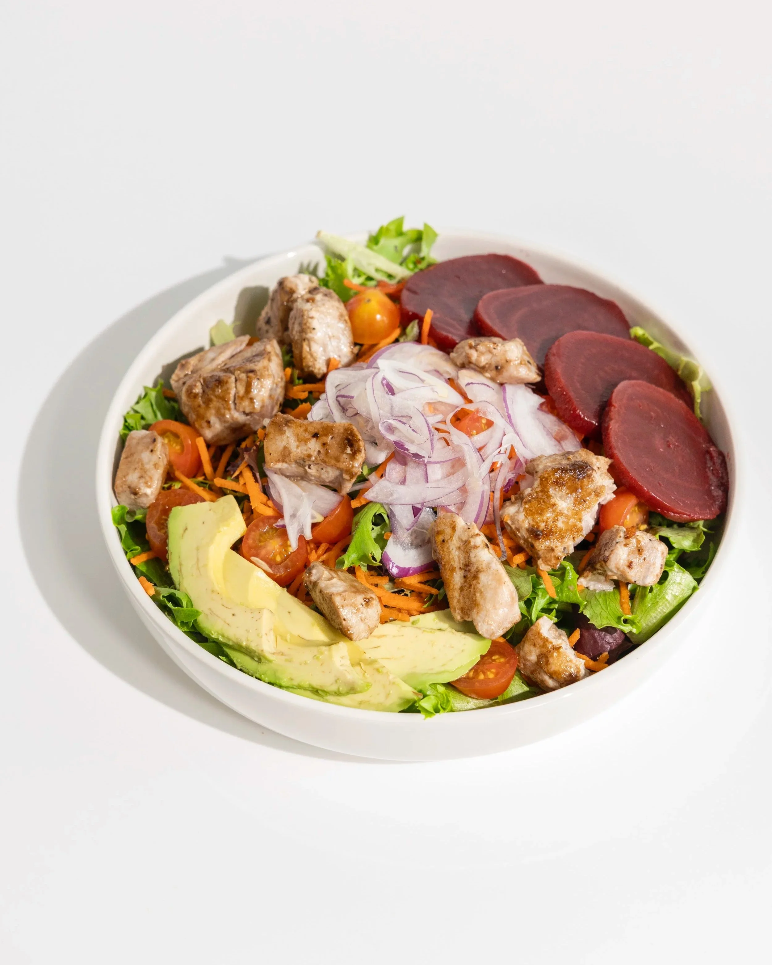 Mixed salad with avocado slices, cherry tomatoes, sliced red onions, shredded carrots, leafy greens, sliced beets, and a side of creamy dressing with black sesame seeds in a small plastic cup