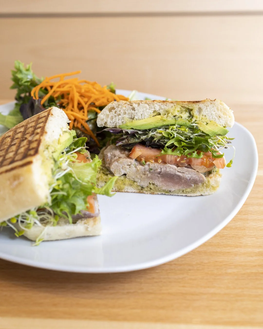 A grilled chicken and vegetable sandwich with lettuce, tomato, avocado, shredded cheese, and mixed greens on a toasted bun, served with a side of shredded carrots and lettuce.
