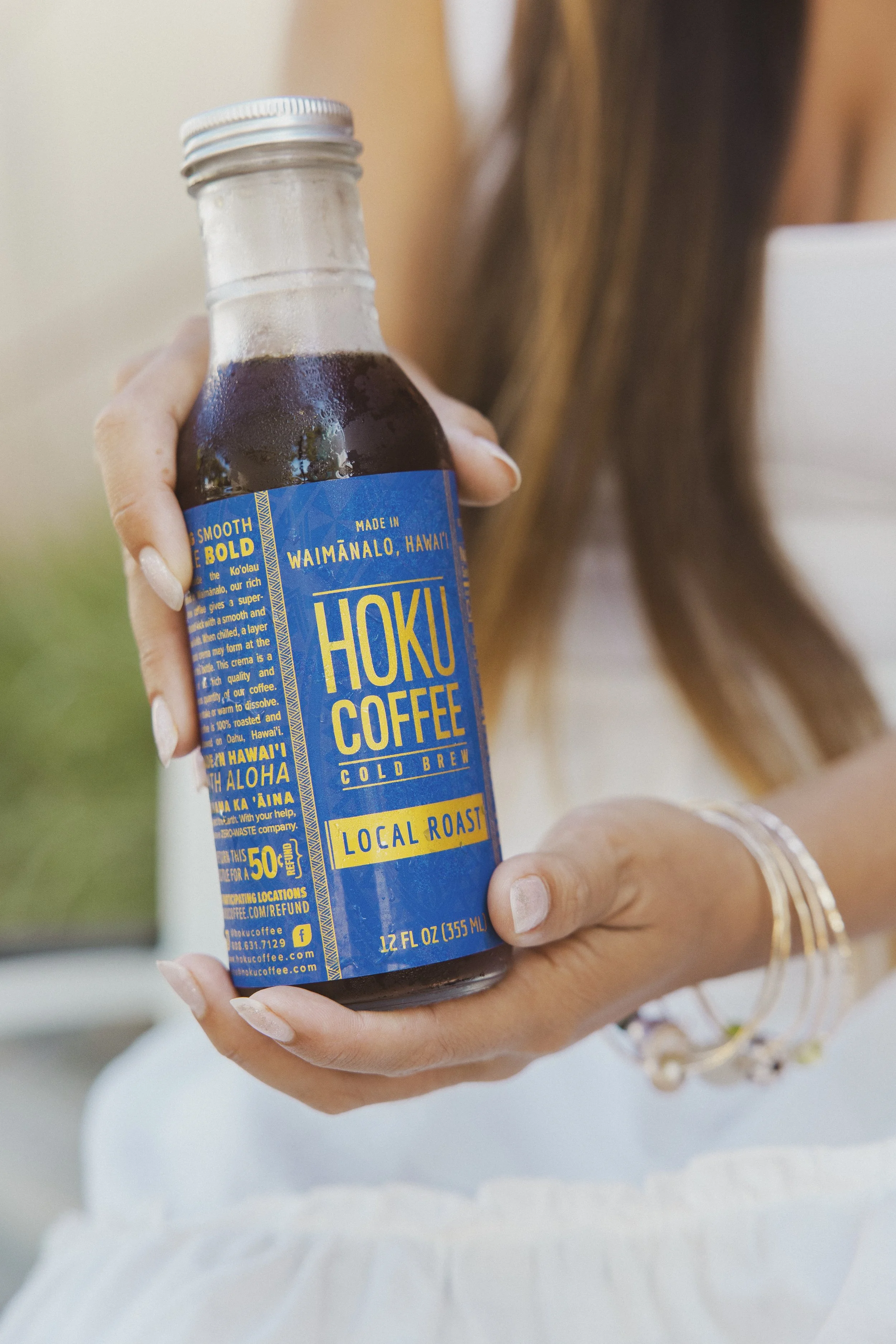 Person holding a bottle of Hoku Coffee cold brew with a blue label, wearing bracelets, with long hair and a white outfit.