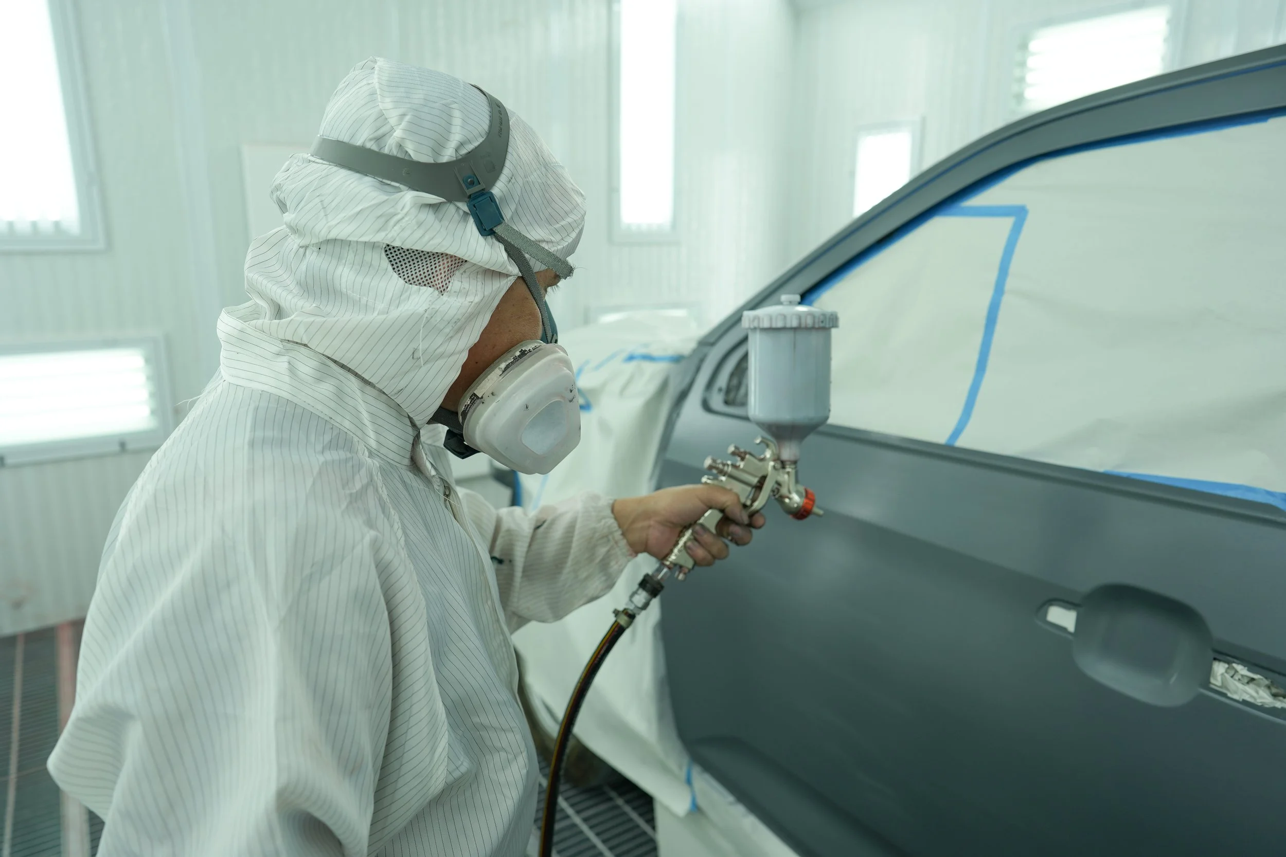 A worker in a cleanroom suit spray-paints a car door in a factory or workshop.