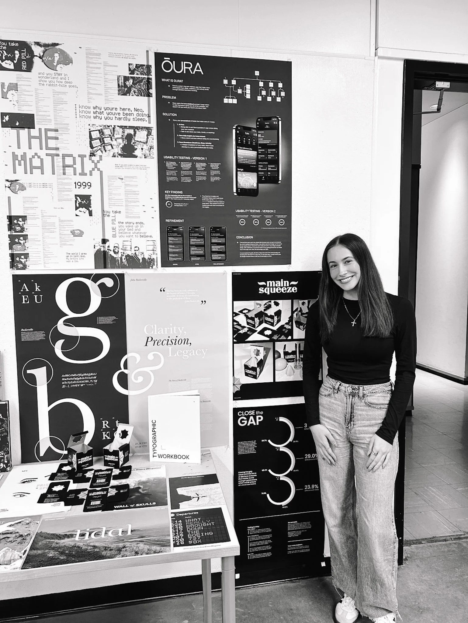 A young woman with long hair standing next to a display board with various posters and graphics, smiling at the camera.