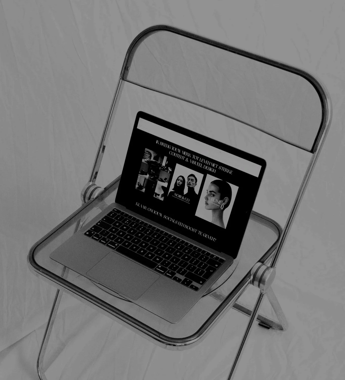Black and white photograph of a laptop on a folding chair with the screen displaying a fashion or magazine website, featuring images of models and text.