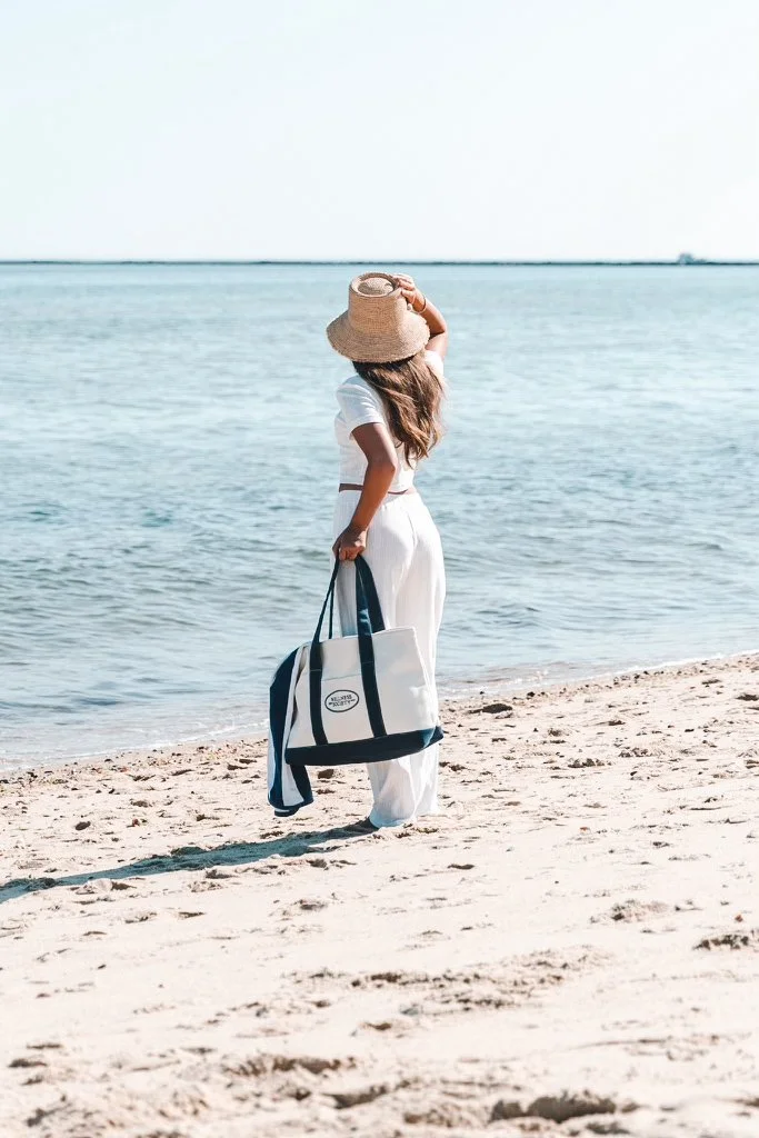 A woman dressed in white standing on a sandy beach near the water, holding a large tote bag and wearing a wide-brimmed straw hat, looking out at the ocean.