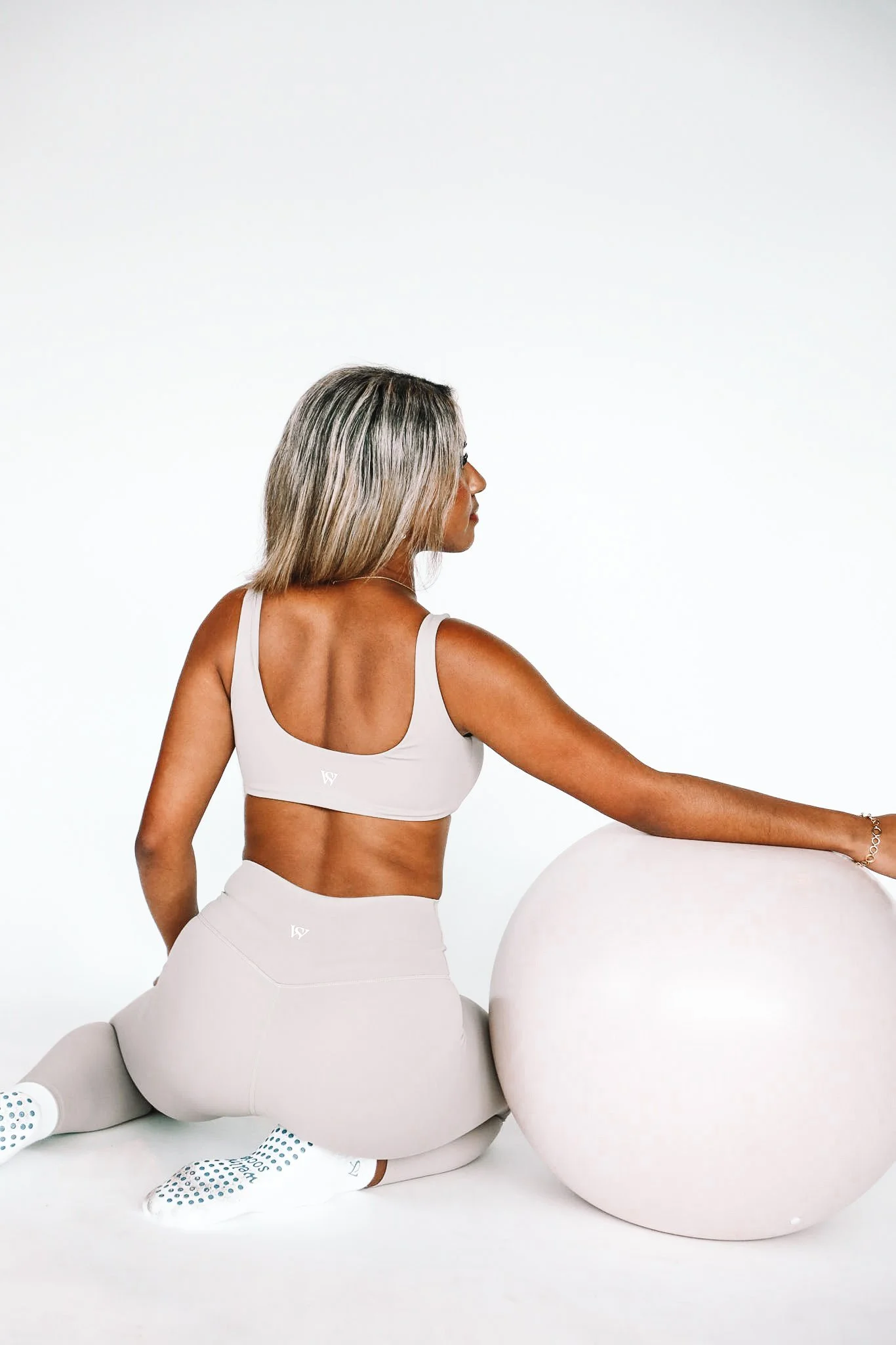 A woman in a beige sports bra, matching leggings, and white socks kneeling on the floor, resting her hand on a large exercise ball against a plain white background.