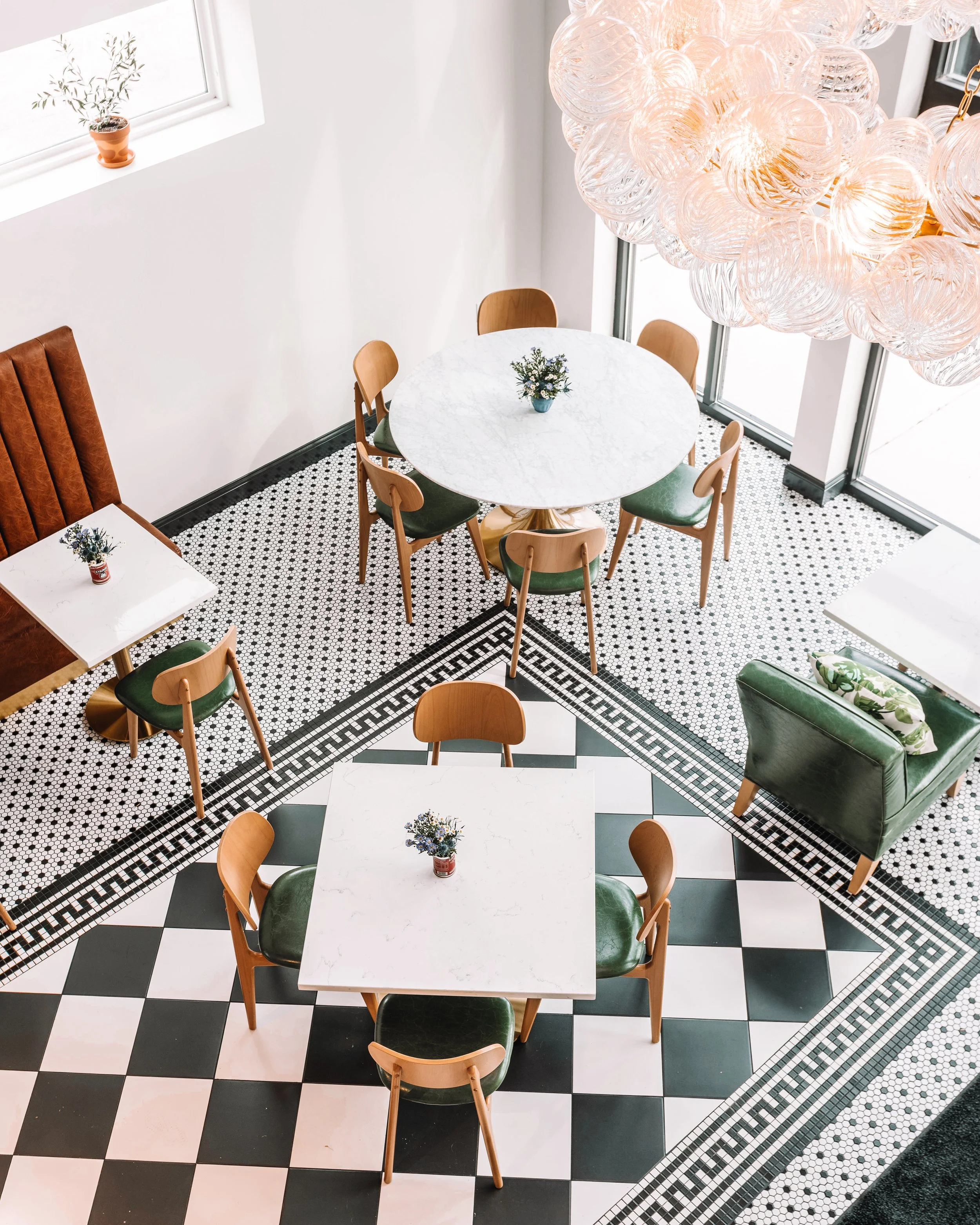 A bright indoor dining area with black and white checkered flooring, a round marble table with wooden chairs, and a green armchair. A unique chandelier made of multiple glass globes hangs from the ceiling. Small flower vases decorate the tables and windowsills.