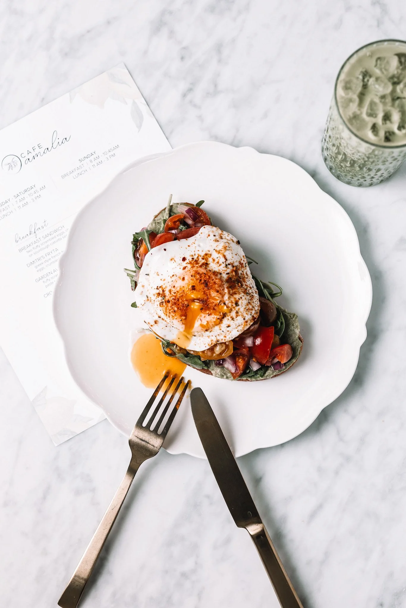 Poached egg on toast with vegetables and a glass of iced beverage on a white marble surface.