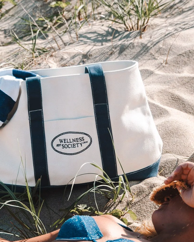 Beach scene with a white and navy tote bag labeled 'Wellness Society', a person with legs and feet visible, lying on the sand, holding a slice of bread or pastry, surrounded by sand and sparse grass.