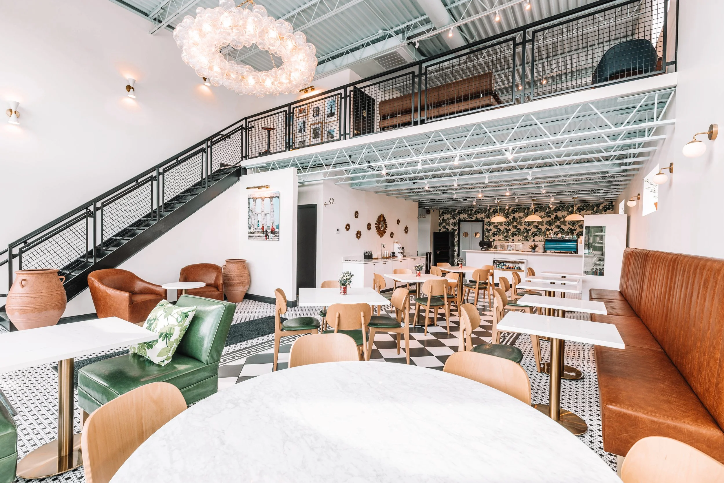 Interior of a modern cafe with white walls, black and white patterned flooring, wooden chairs, green and brown leather seating, white tables, a decorative floral wall, and a staircase with black railings leading to an upper level, with visible lighting fixtures and a chandelier.