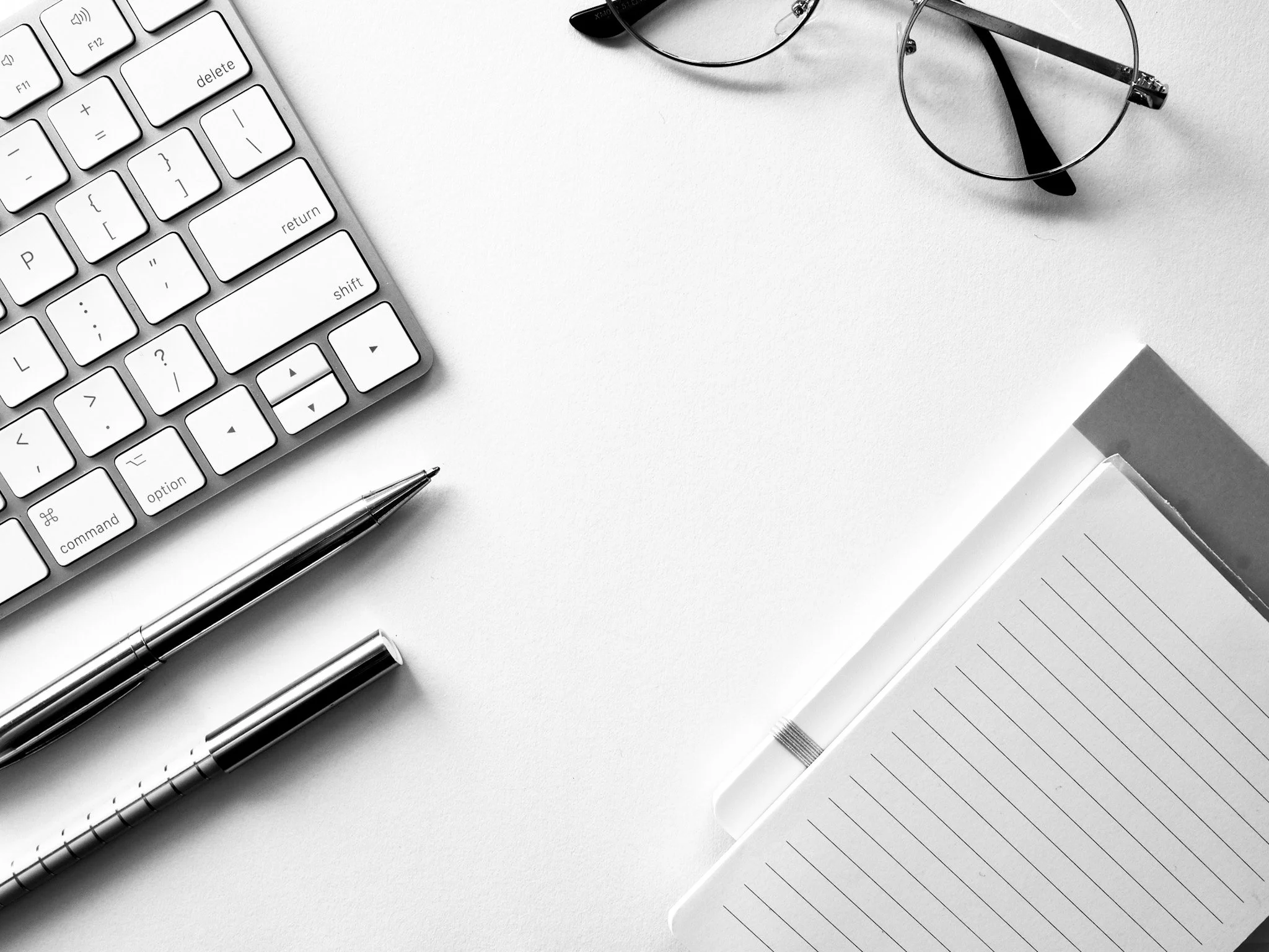 Black and white photo of a neatly organized workspace with a computer keyboard, a pair of glasses, two pens, and an open notebook on a white surface.