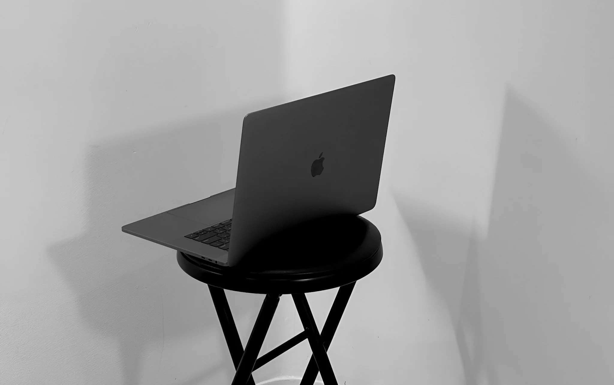 A black MacBook laptop on a round black stool against a plain white wall.