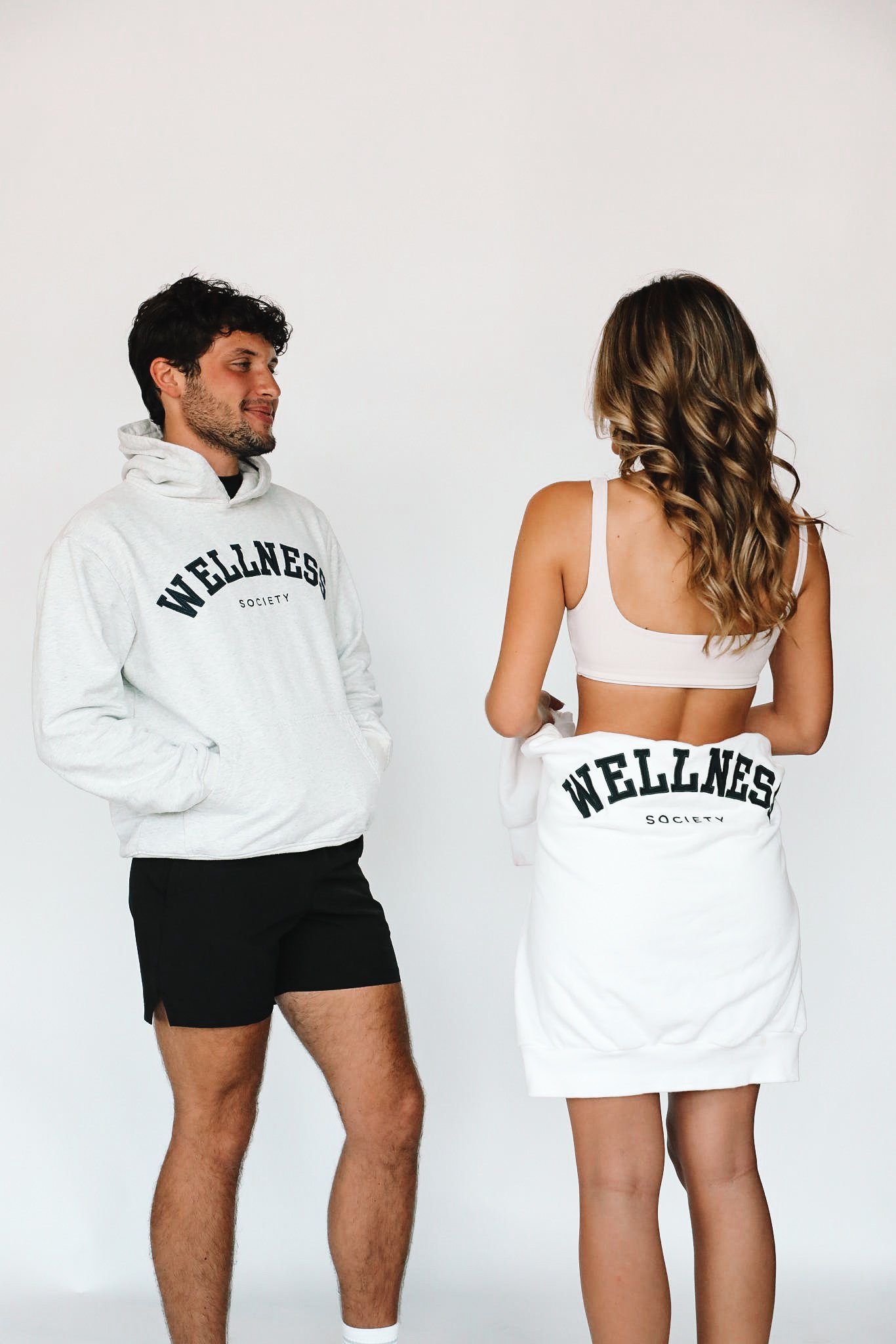 A man and woman are standing and facing each other, both wearing matching white clothing with 'WELLNESS' printed on it. The man is in a hoodie and shorts, while the woman is in a sports bra and a towel or blanket with the same text.