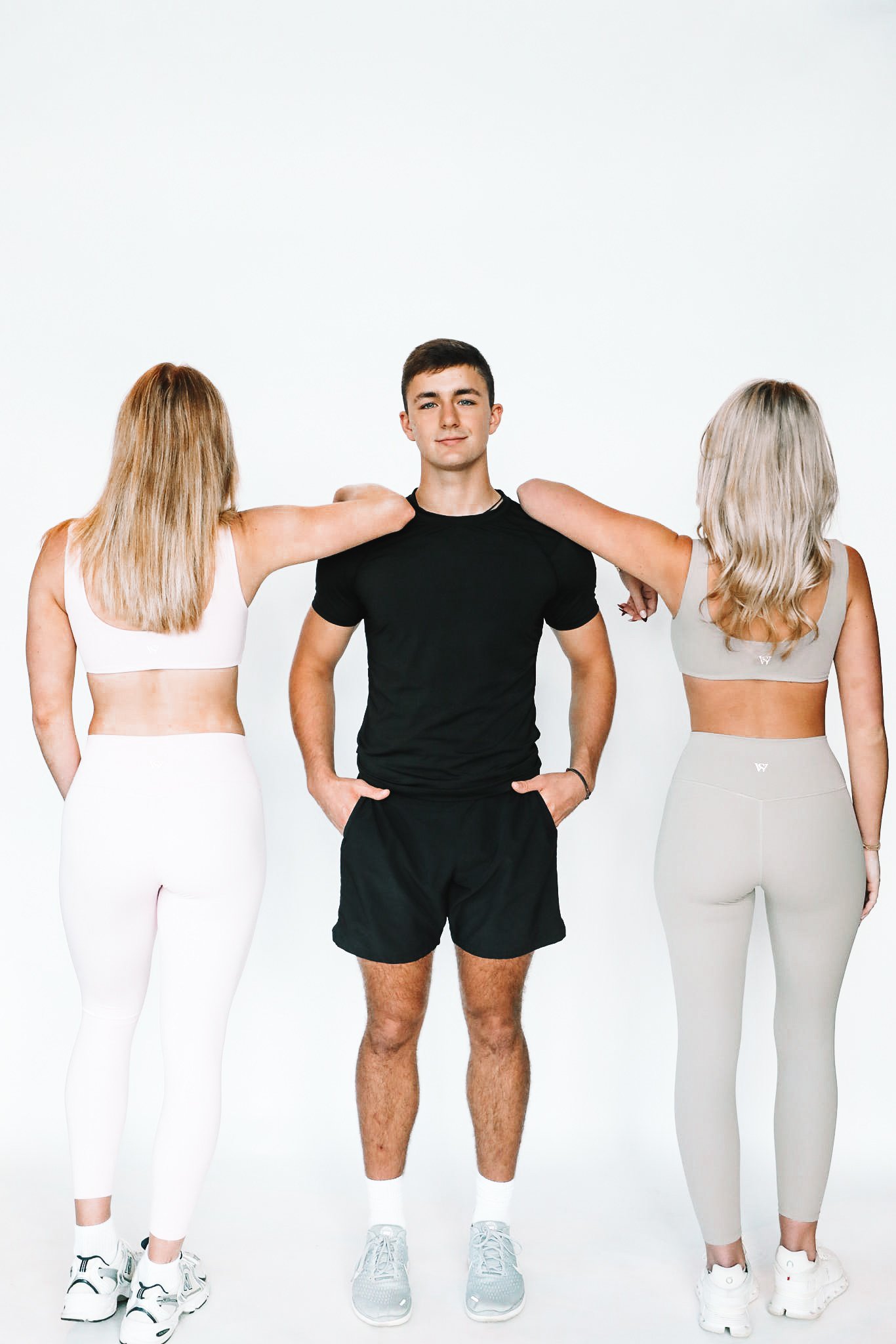 Young man in black athletic outfit standing between two women in workout clothes with their arms on his shoulders in a fitness studio with a white background.