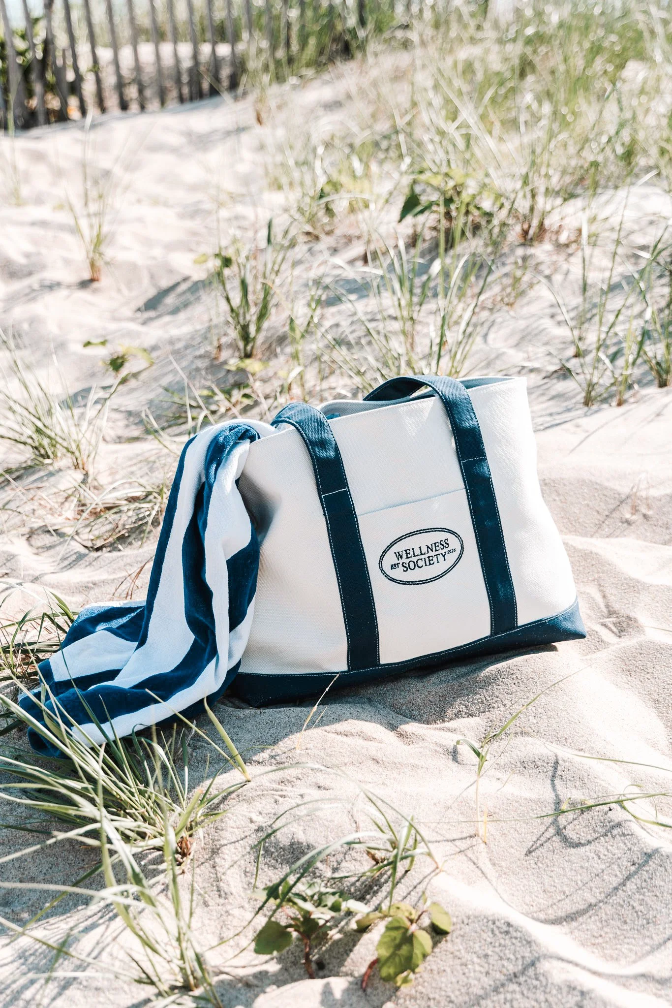 White and navy tote bag with the words 'Wellness Society' on a sandy beach with grass and a railing in the background.