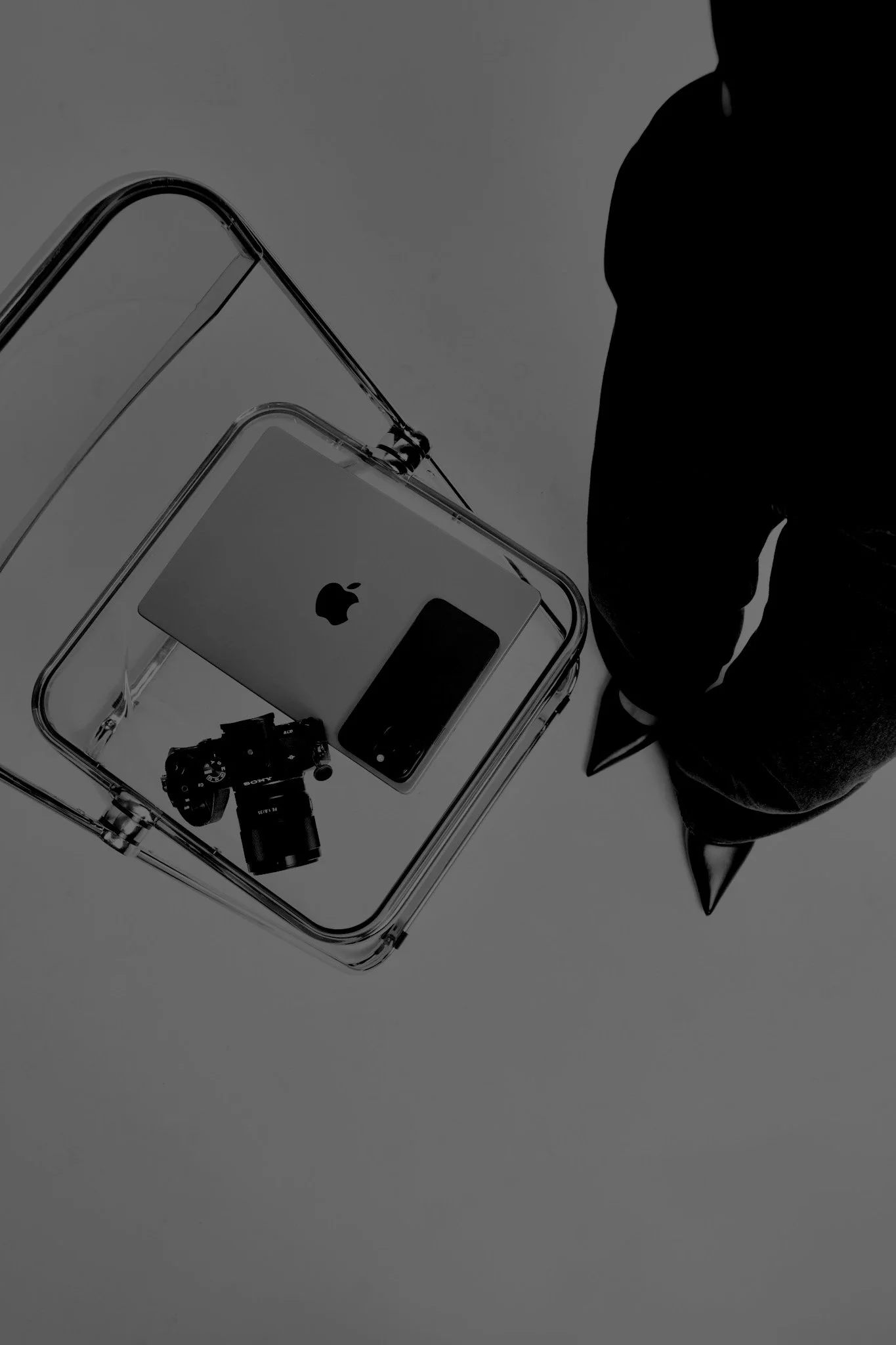 Top view of a glass display case with a white iPhone, a small camera, and a black object inside it, next to a person wearing a dark jacket and black pointed shoes.