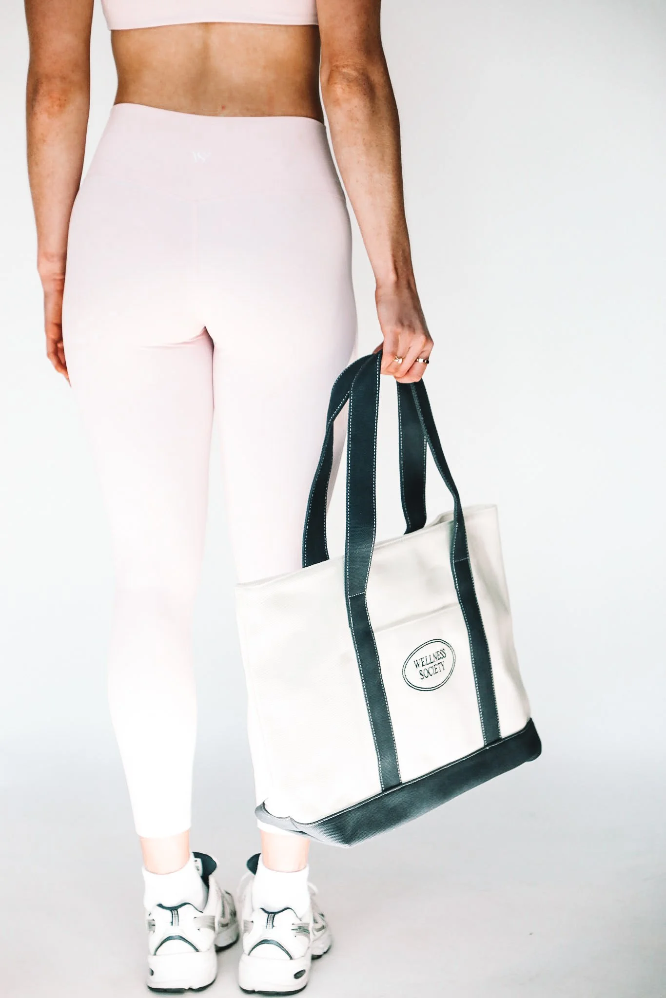 Person in pink athletic clothing standing against a plain backdrop, holding a white tote bag with black straps that reads "Wellness Society."