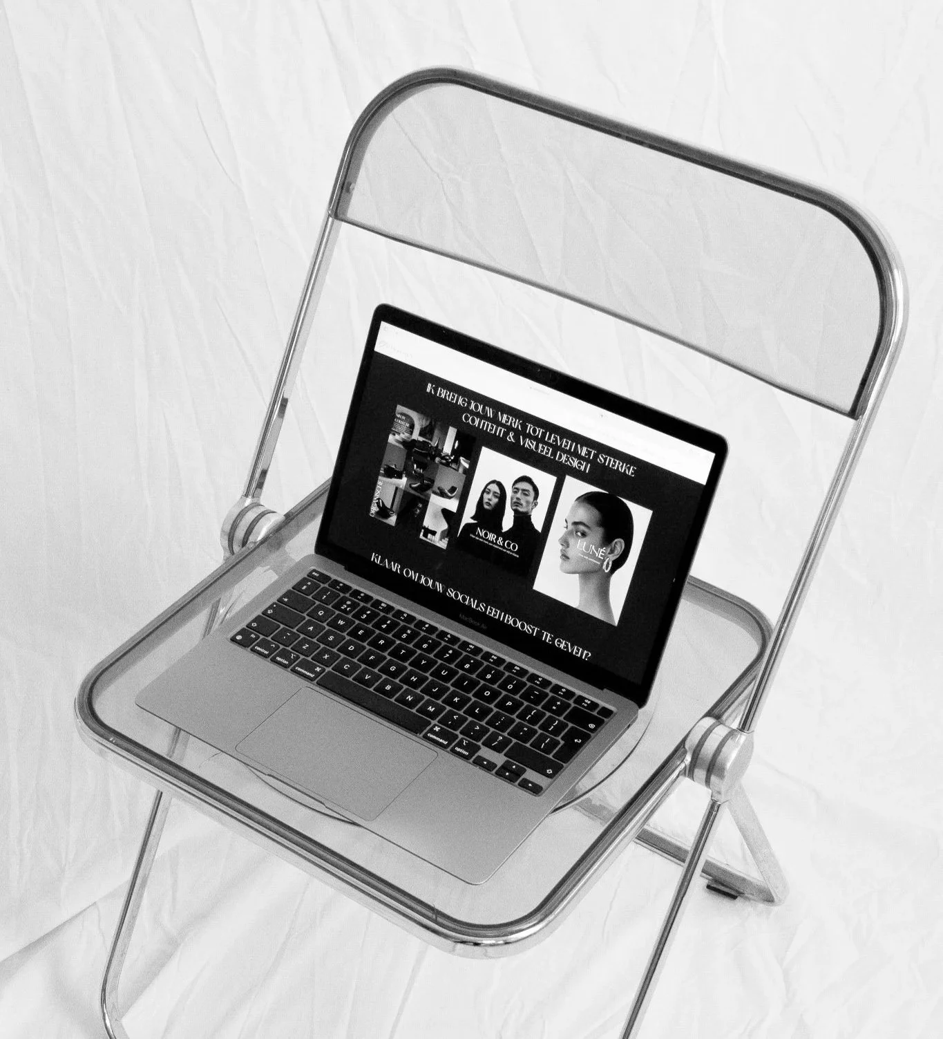 A laptop on a clear plastic chair displaying a webpage with black and white images of models and text in a non-English language.