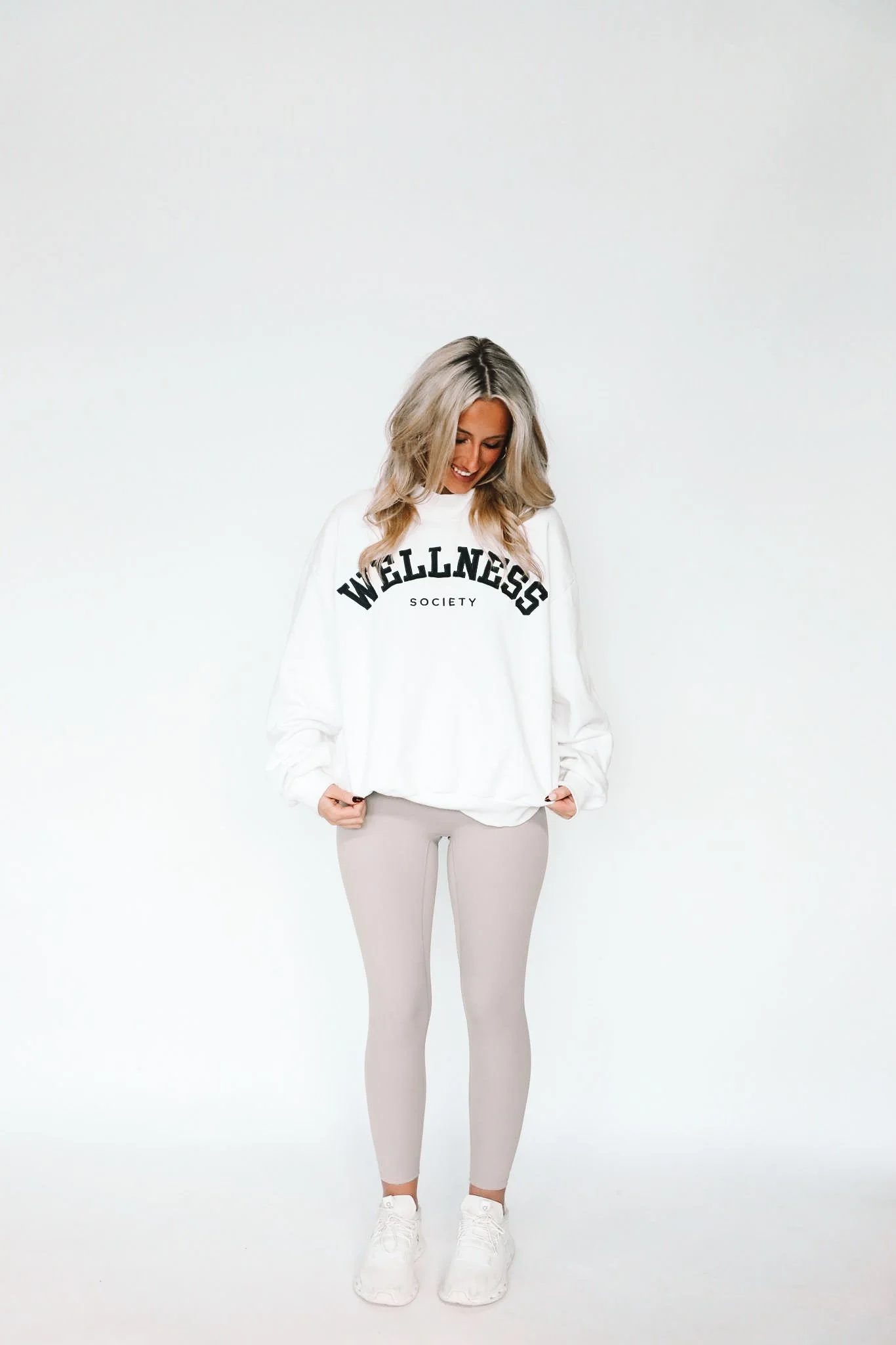 Woman with long blonde hair standing against a plain white background, wearing a white sweatshirt with 'WELLNESS SOCIETY' written on it, light gray leggings, and white sneakers. She is looking down and smiling.