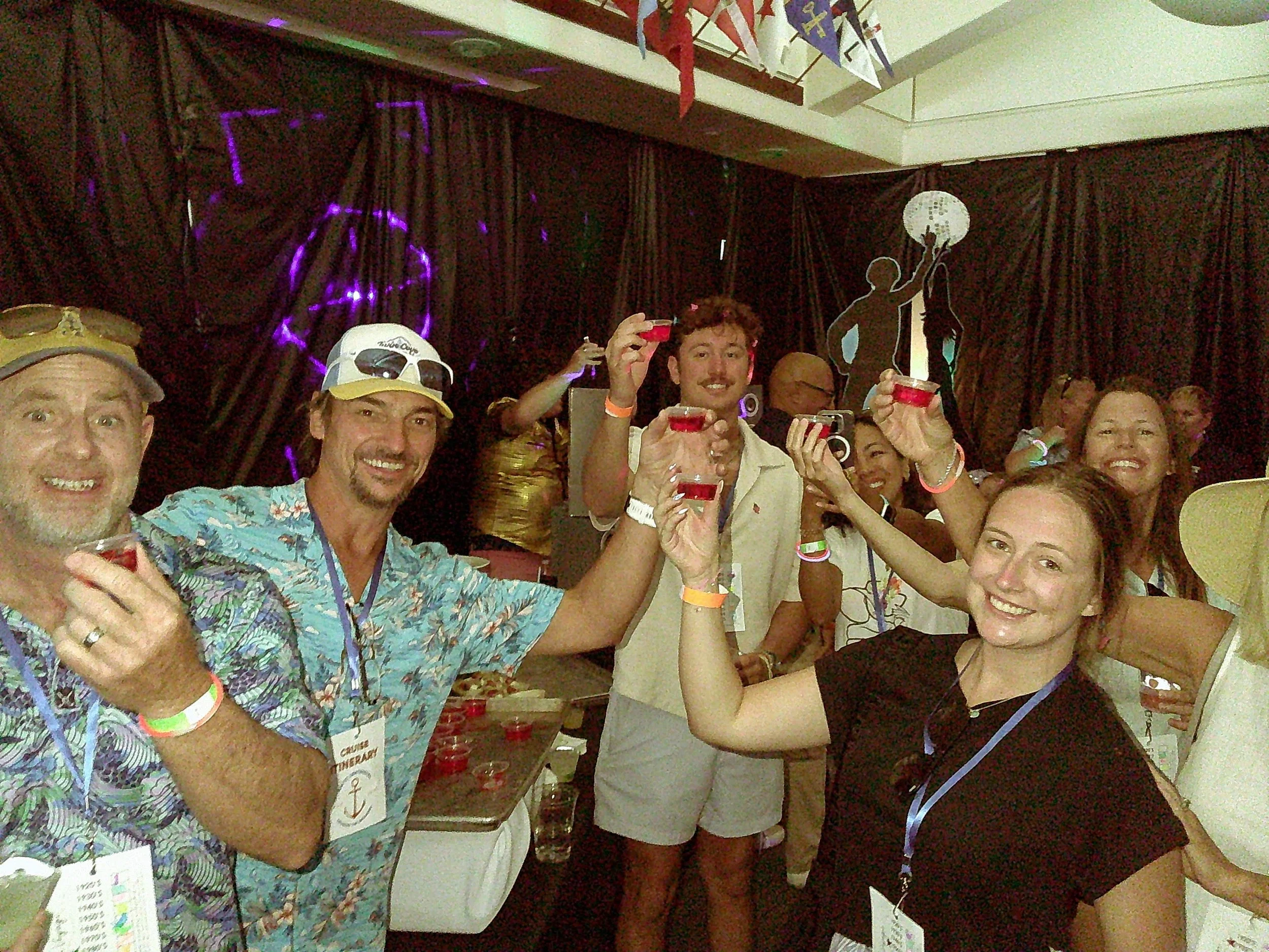 Group of people at a party or celebration raising small cups of red drink in a toast, smiling and enjoying the event, with a dark curtain backdrop and colorful decorations.