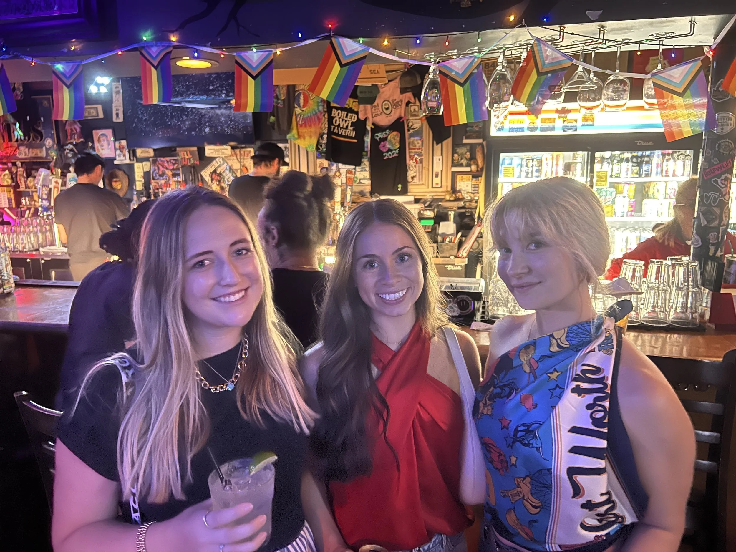 Three women enjoying drinks at a lively bar decorated with rainbow pride flags, with colorful lights and memorabilia in the background.