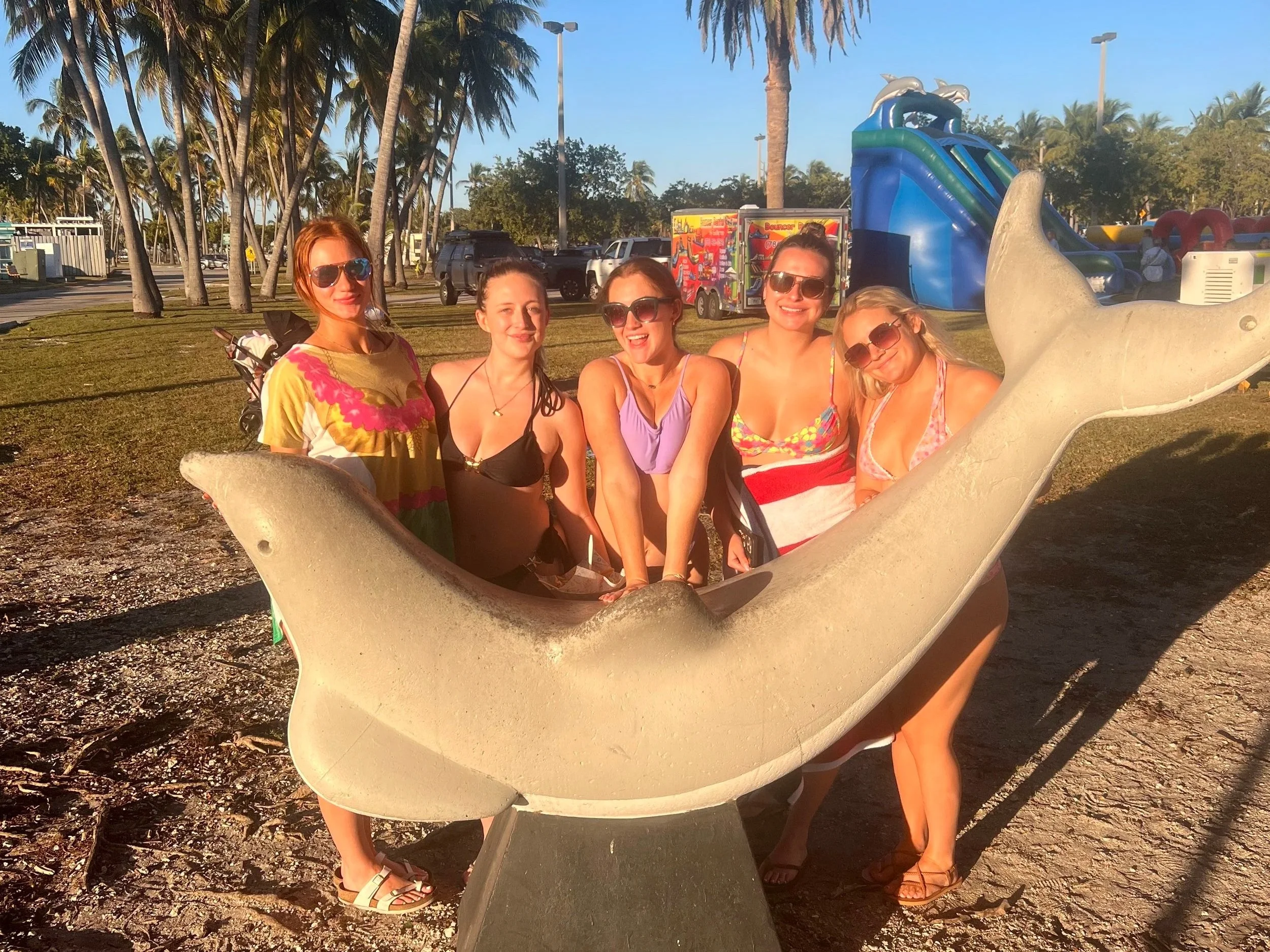 Five women in swimsuits and sunglasses posing around a dolphin sculpture at a water park or beach area with palm trees, kiddie slides, and bright blue sky in the background.