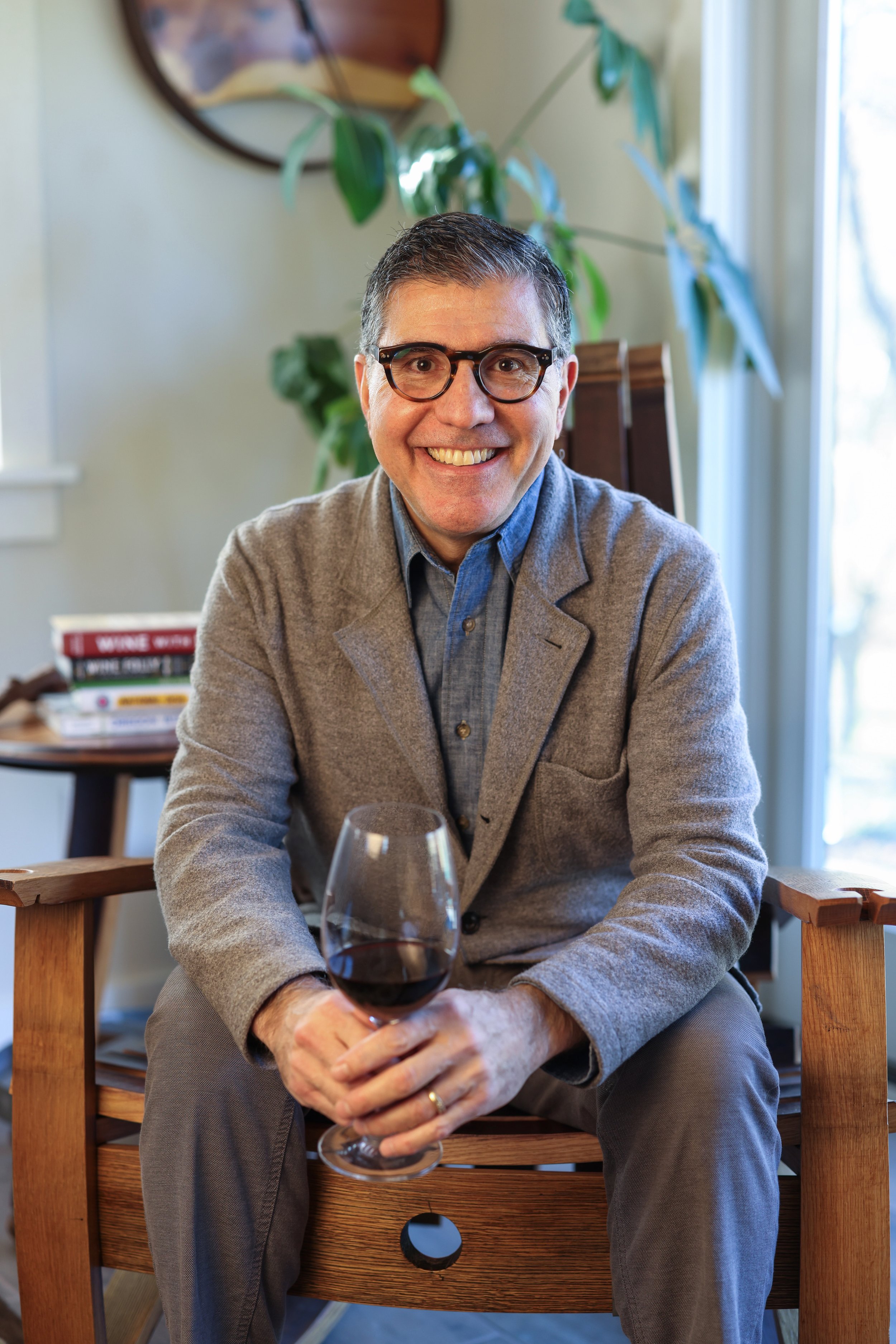 Man with glasses and a gray blazer smiling and holding a glass of red wine while sitting on a wooden chair in a cozy room with a bookshelf and houseplant in the background.