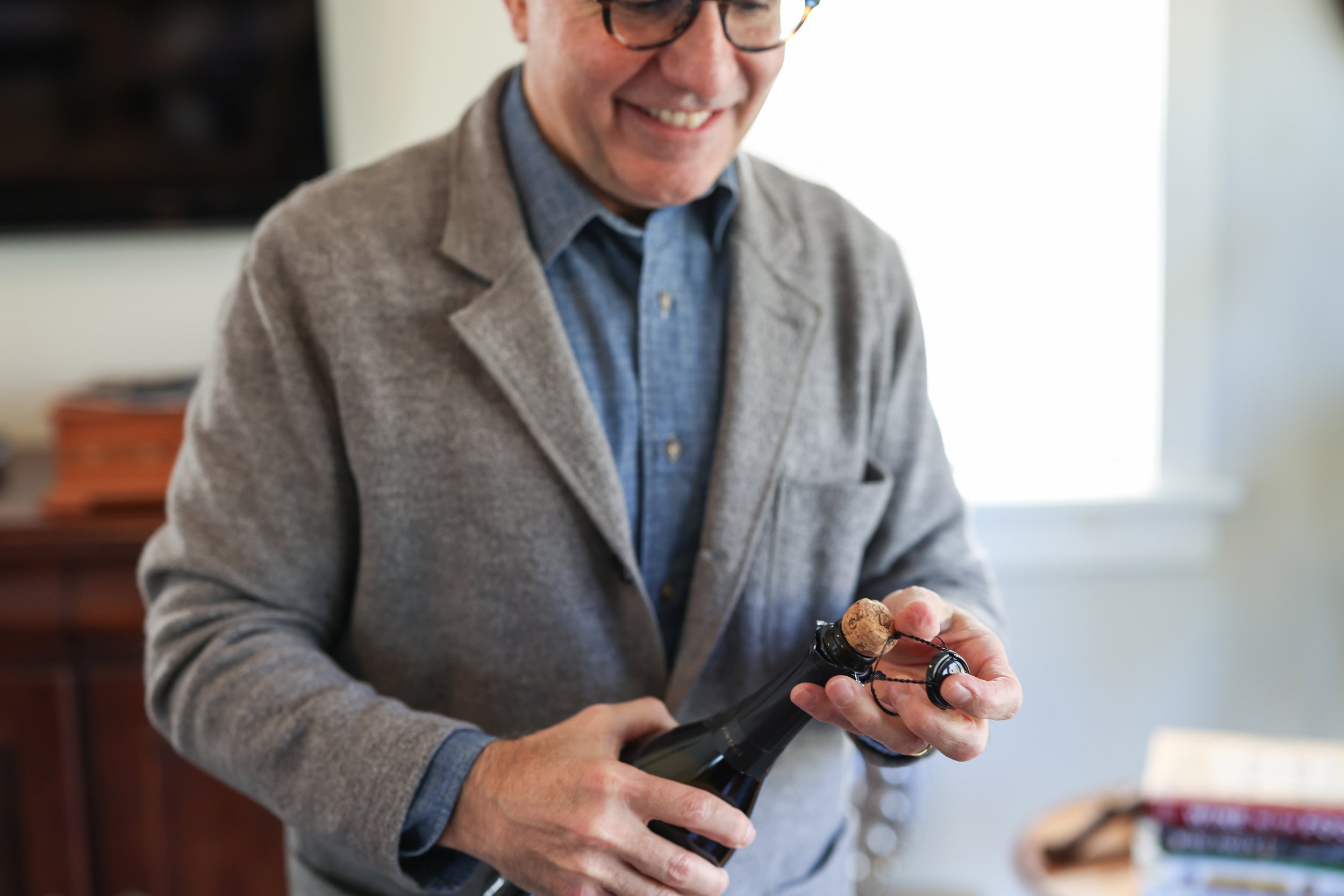 A man is opening a bottle of champagne with a corkscrew, smiling and wearing glasses, a gray jacket, and a blue shirt, in a bright room.