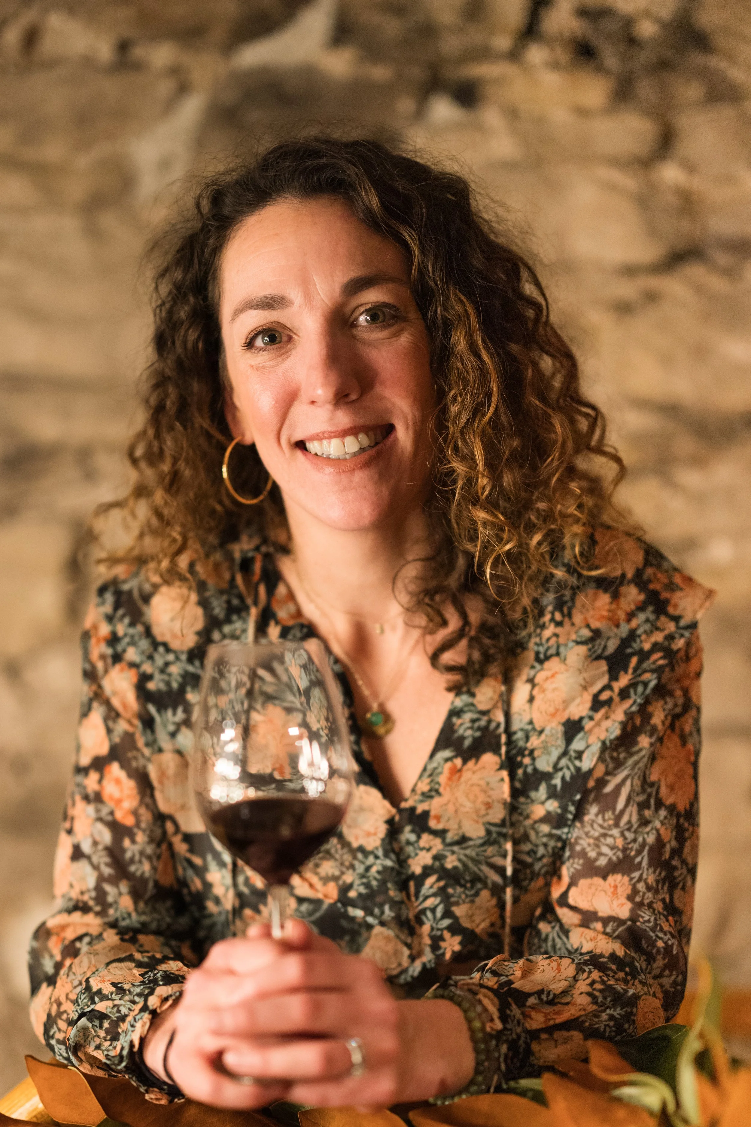 A woman with curly brown hair and hoop earrings smiling at the camera while holding a glass of red wine, sitting in front of a stone wall.