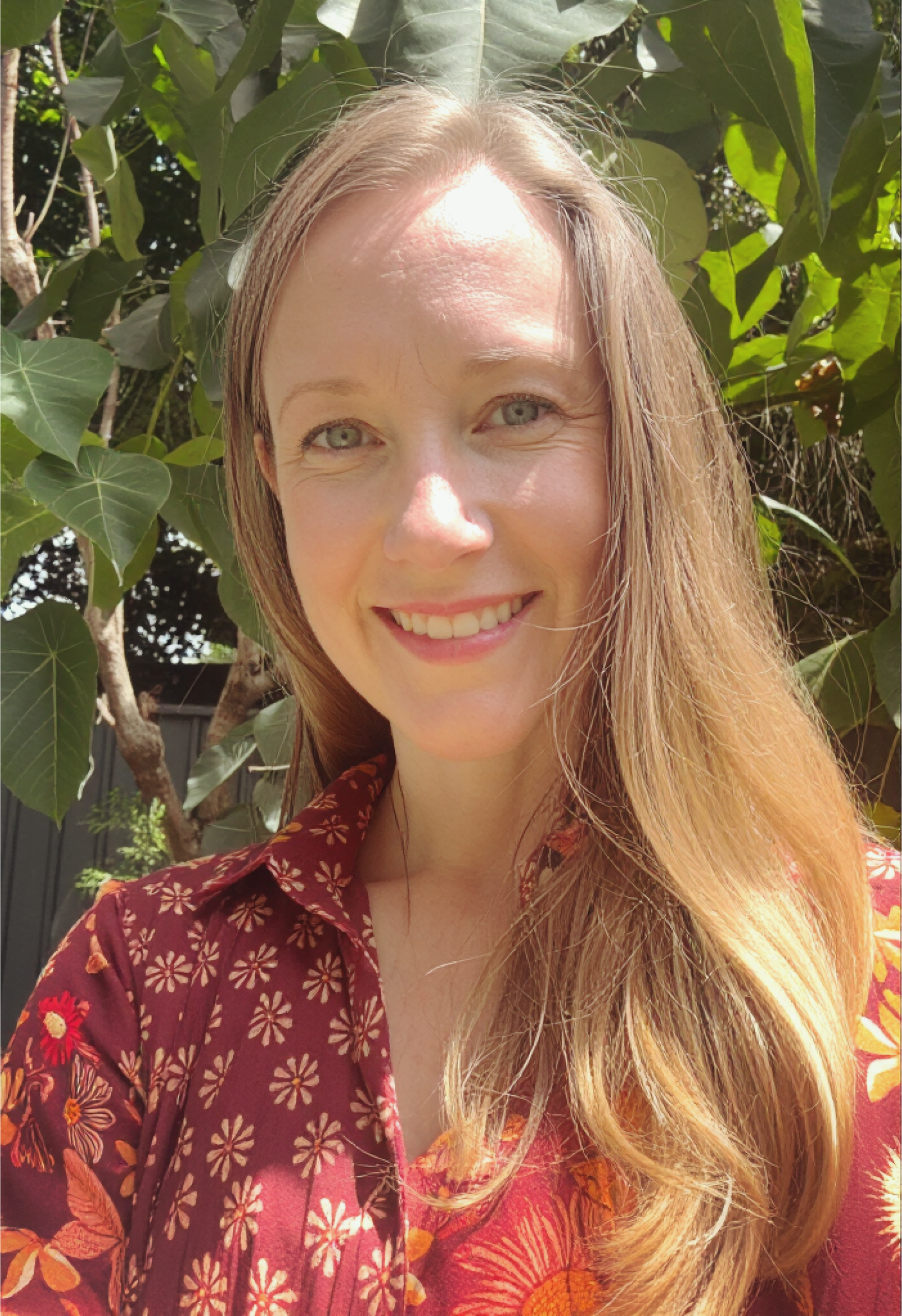 A smiling woman with blonde hair, wearing a blue floral dress, sitting indoors in front of light-colored walls.