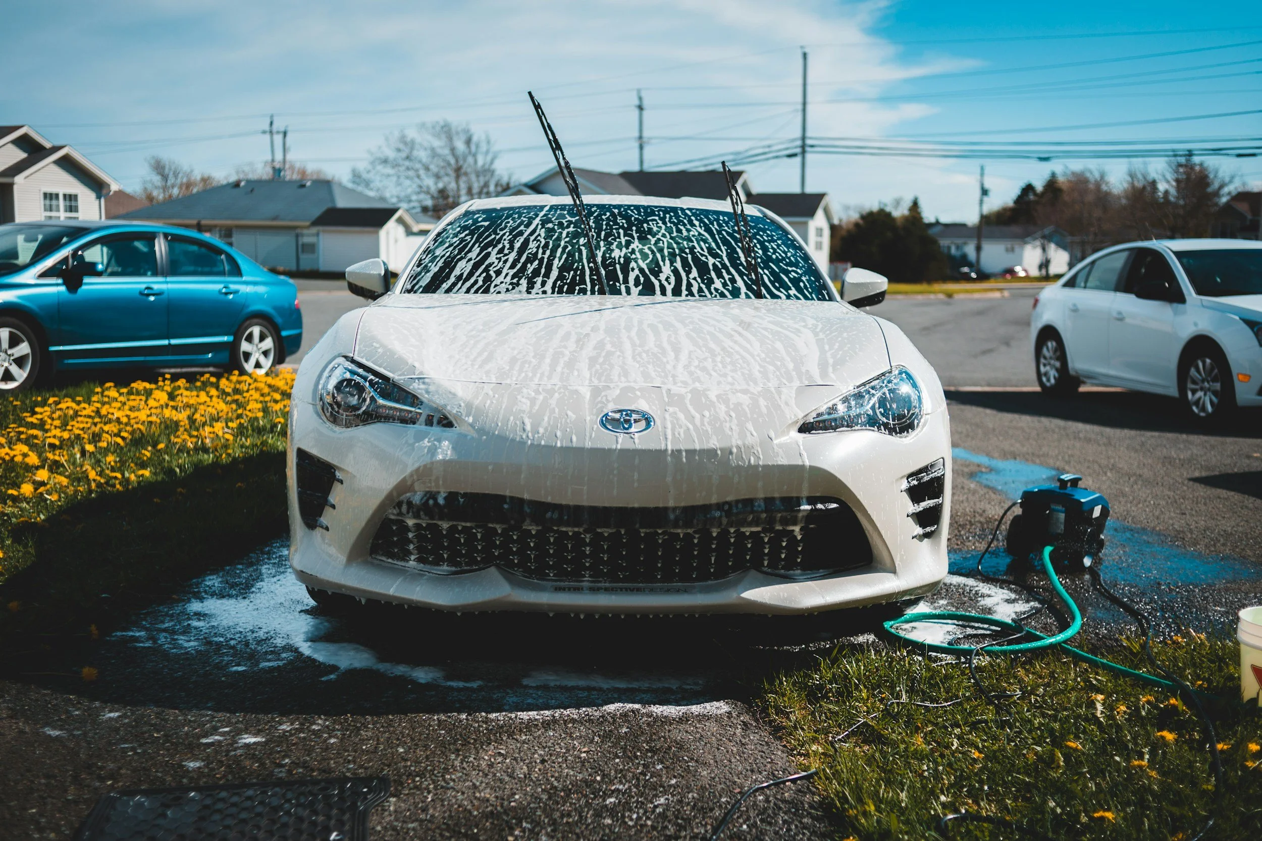 White sports car being washed with soap suds on the front, in an outdoor car wash area with other parked cars and residential houses in the background.