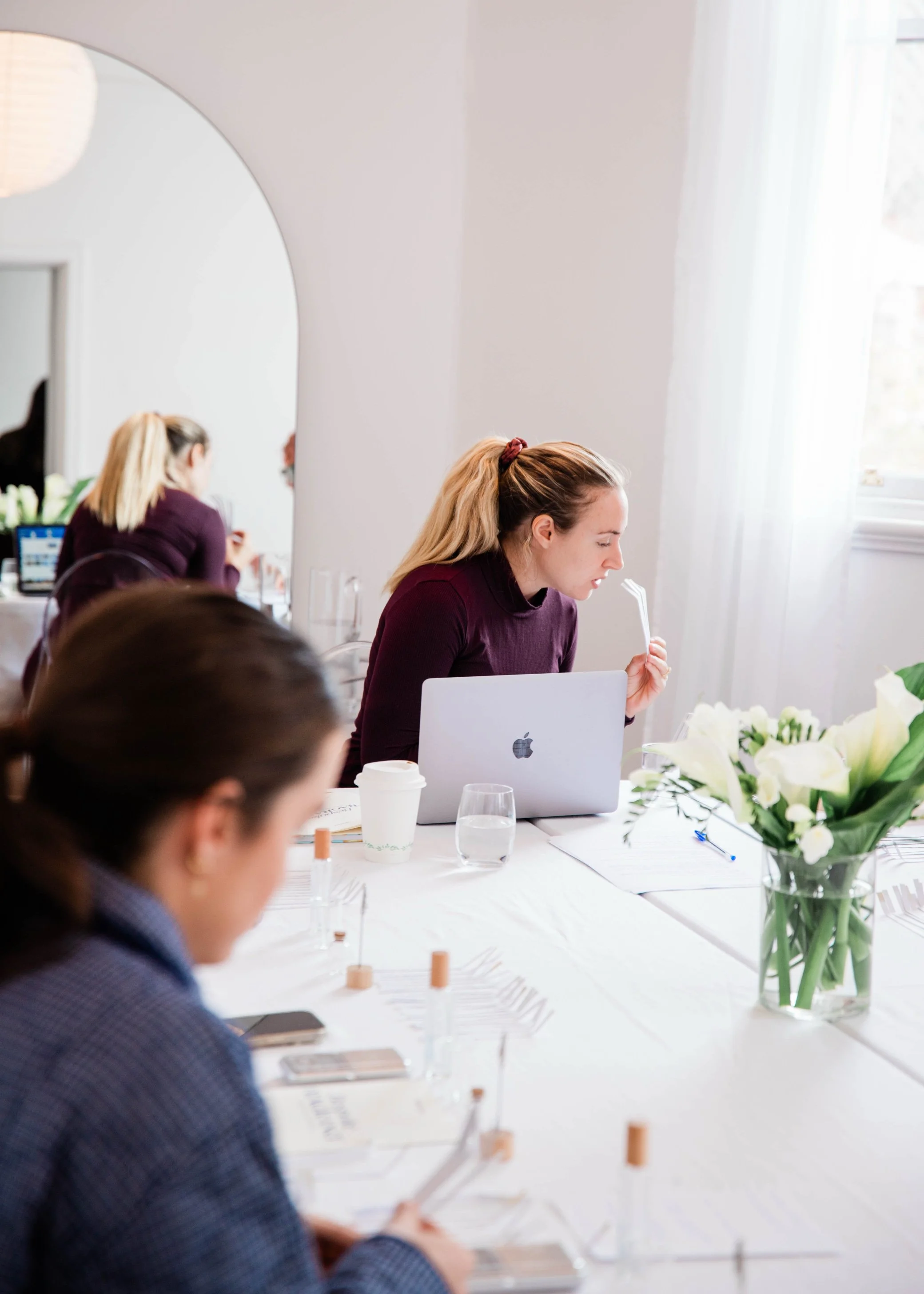A woman in a maroon top sitting at a desk, eating with a fork. She has a laptop, a glass of water, and a to-go coffee cup in front of her. There are white flowers in a vase on the table, and another person in the foreground is reading or writing.