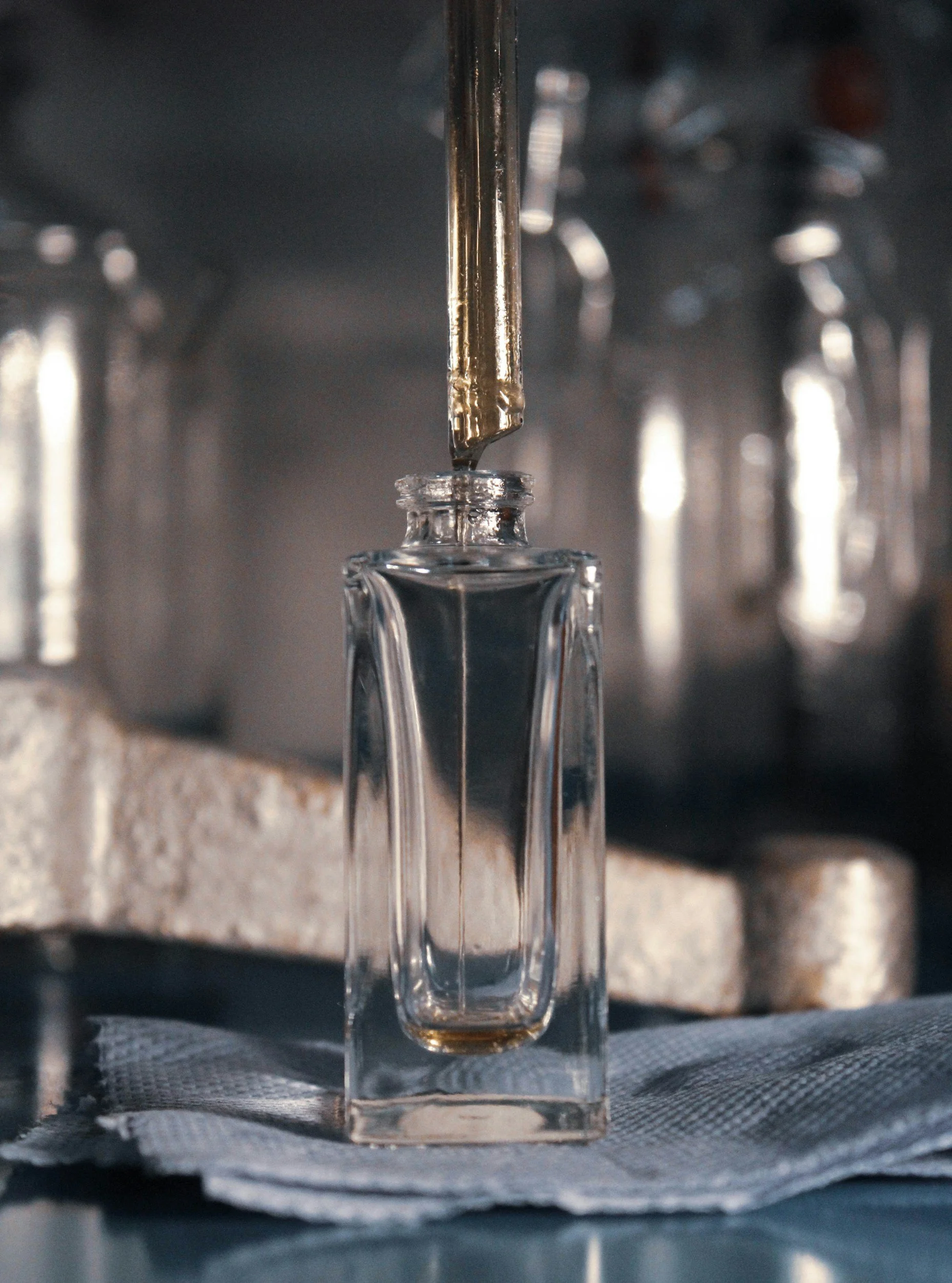 A glass perfume bottle being filled with perfume from a dropper, with a blurred background of metallic objects.