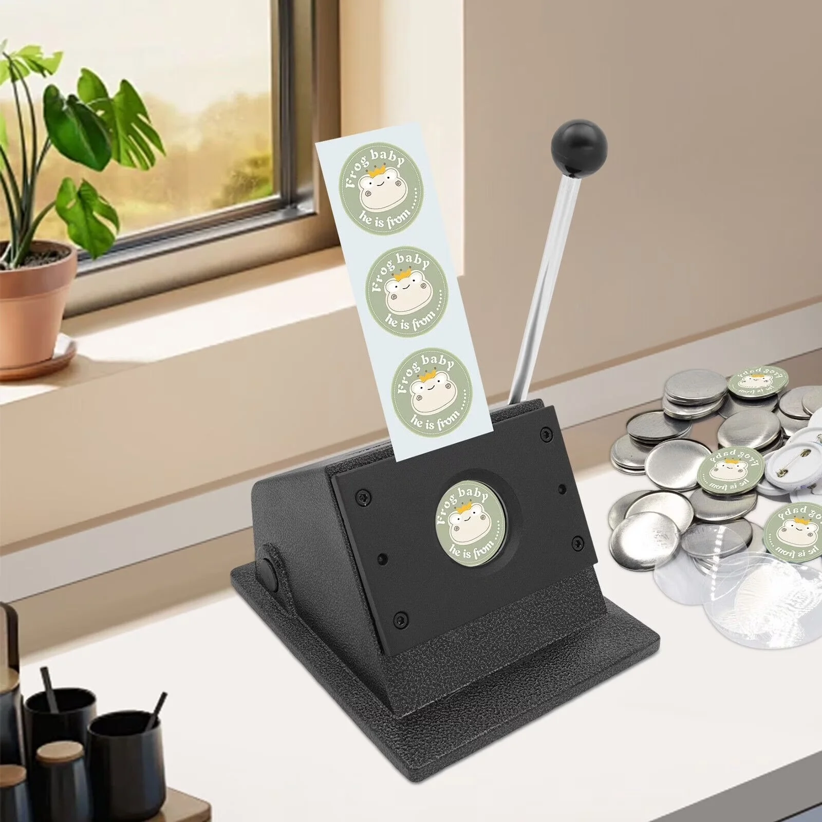 A badge maker machine on a white table printing stickers featuring a cute cartoon hippopotamus with a crown, the stickers read 'Frog's baby he is from'. Several printed stickers and blank metal backing circles are scattered on the table nearby, with a potted plant and a window in the background.